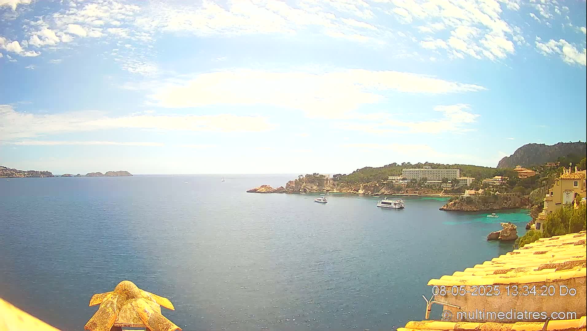 A partly sunny day reveals a coastal scene with calm blue water, rocky shores, buildings, and boats, viewed from an elevated vantage point.