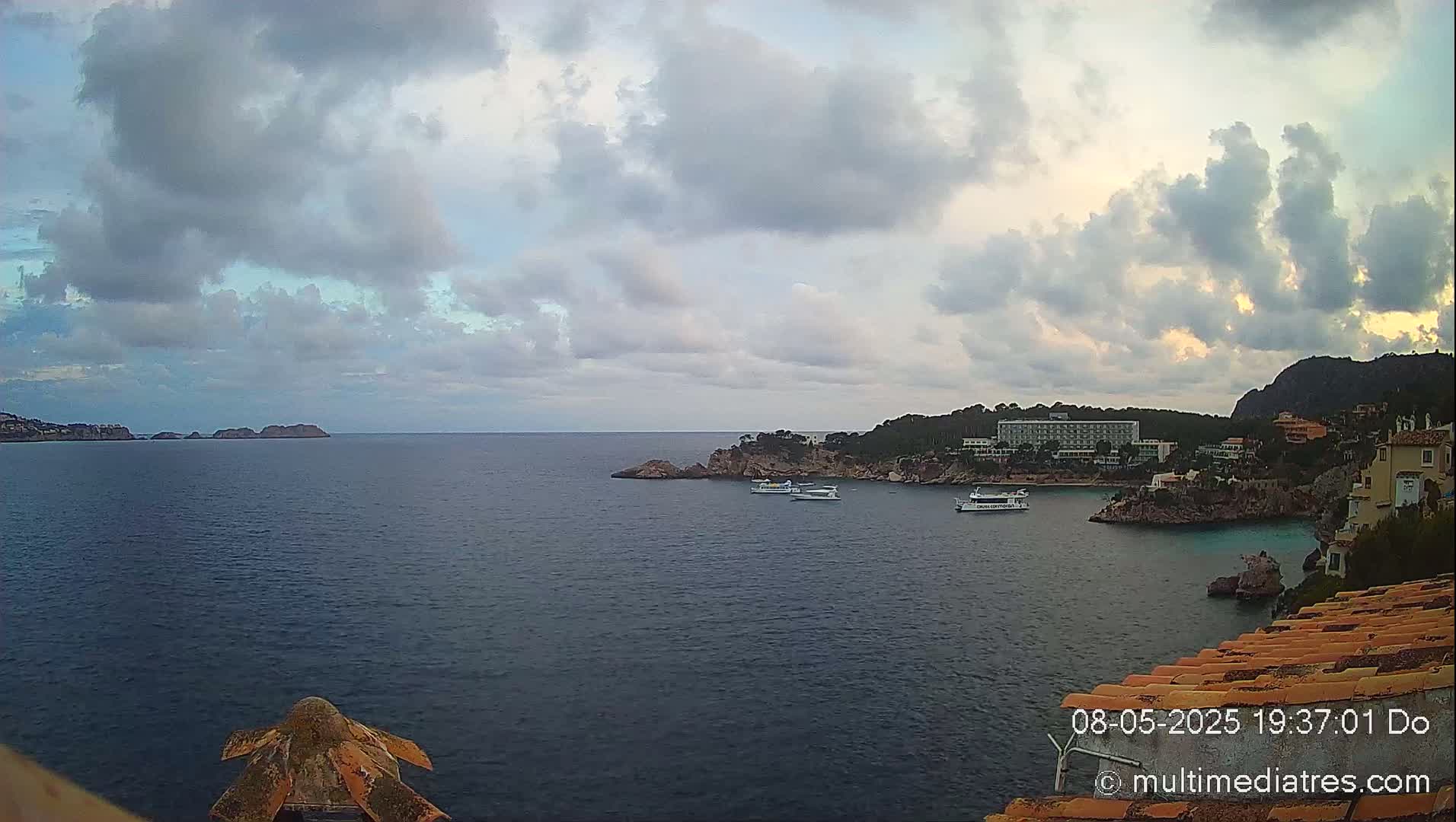 A partly cloudy evening overlooks a calm bay with several boats and a hotel nestled into a rocky coastline.