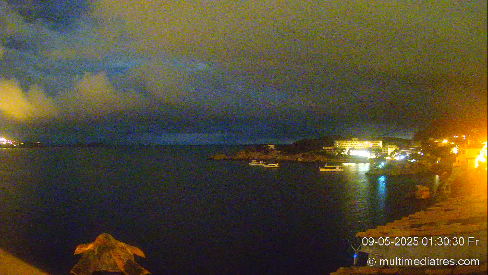 A nighttime view of a calm body of water with a few boats and illuminated buildings on the shore under a cloudy sky.