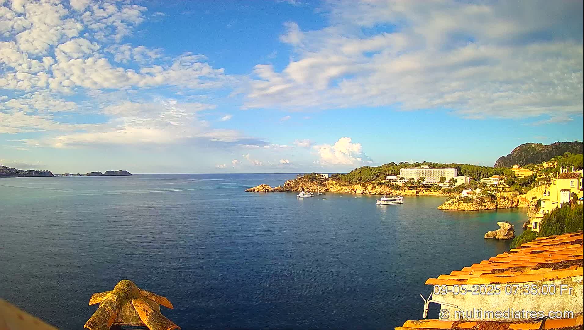 A partly cloudy sunny day reveals a tranquil bay with calm water, a few boats, and buildings along a rocky coastline.
