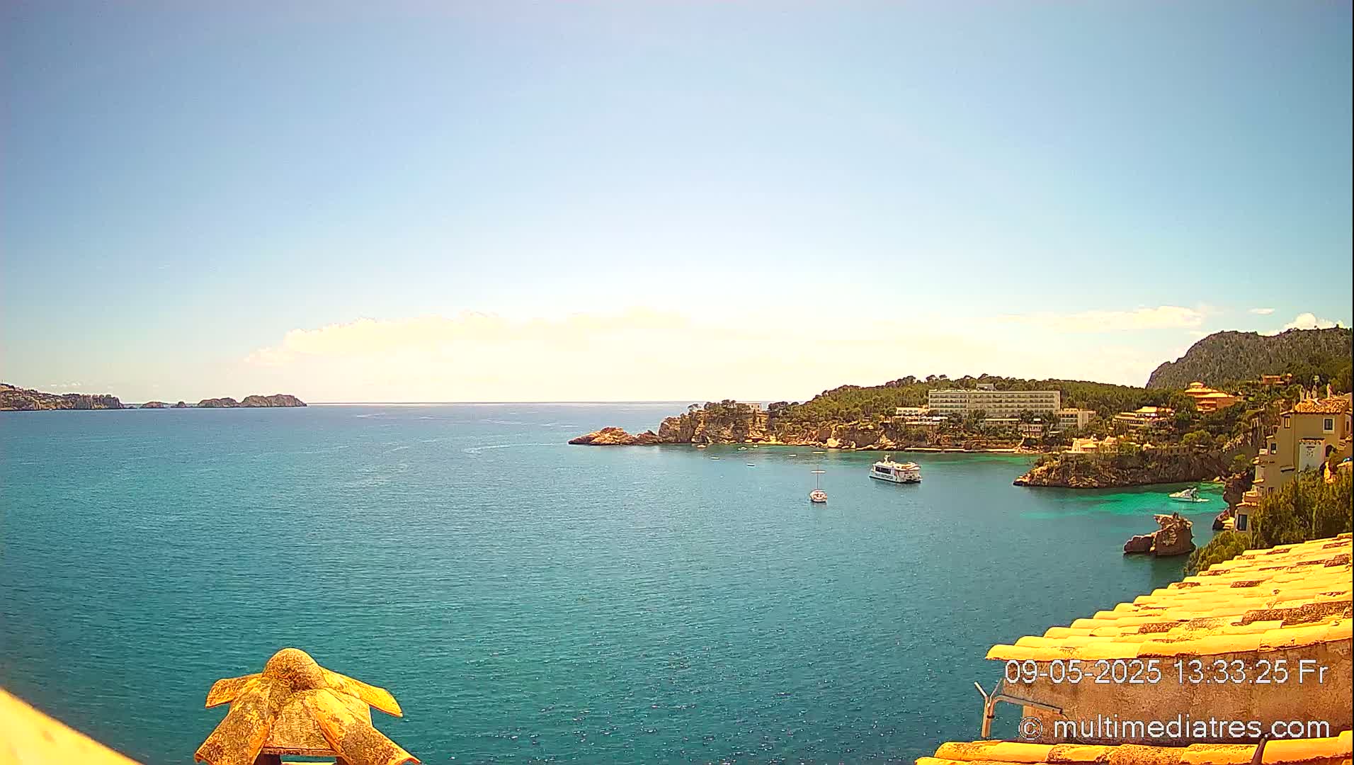 A sunny, bright day shows a coastal scene with teal water, rocky cliffs, buildings, and boats.