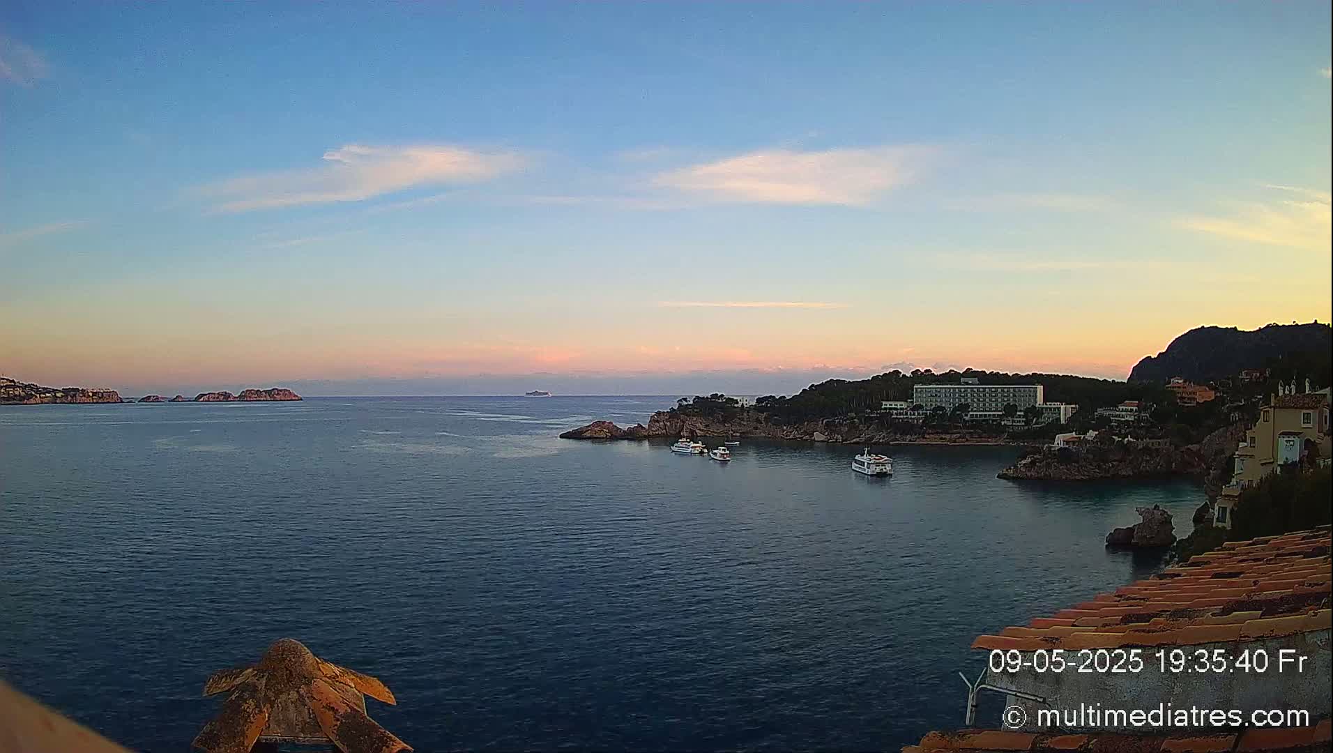 A tranquil coastal scene at sunset shows a calm sea with several boats near a rocky shoreline, featuring a large hotel and distant islands under a clear, pale sky.