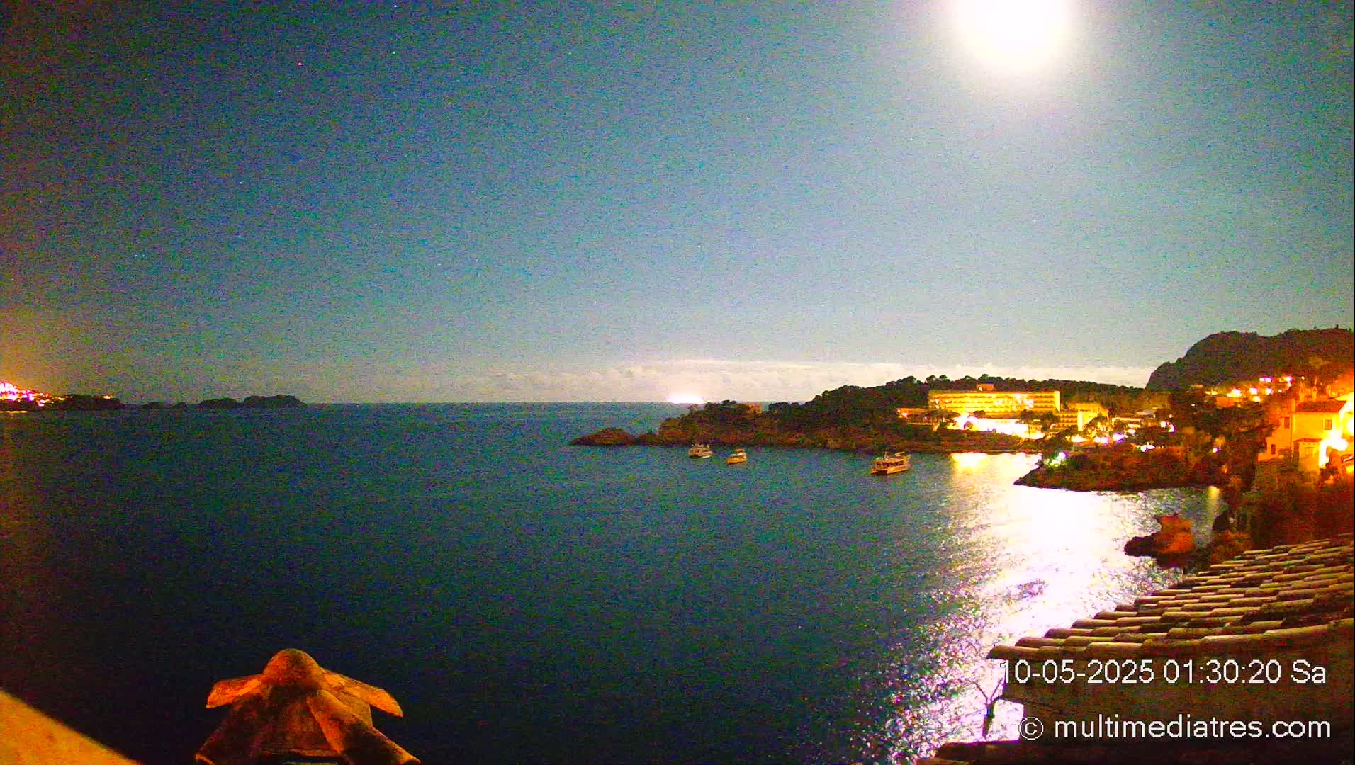 A moonlit nighttime scene shows a calm body of water with a few boats, a shoreline with buildings and lights, and a dark hill in the background under a clear, starry sky.