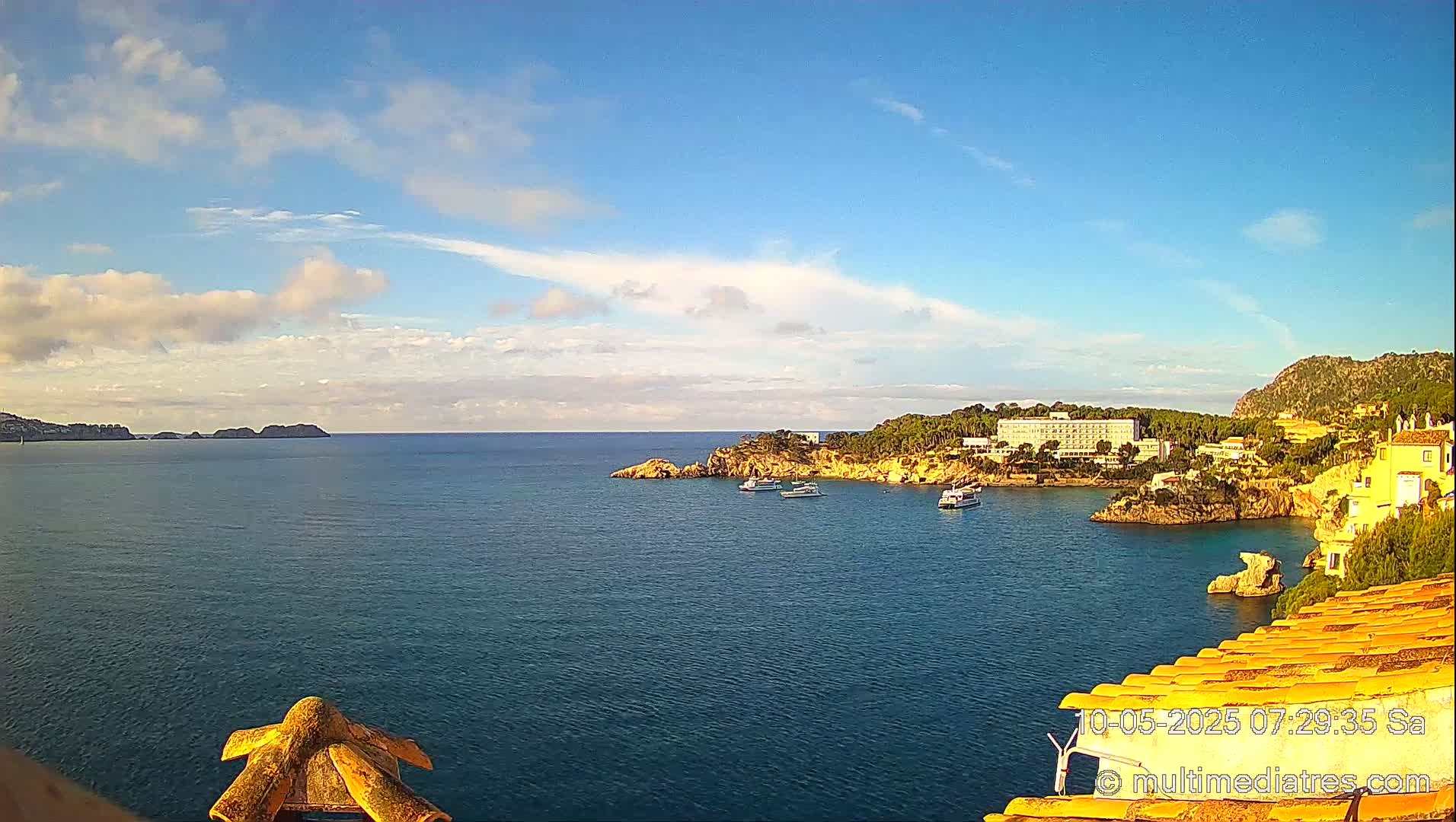 A calm, blue sea with a few boats is seen from a high vantage point, under a mostly sunny sky with scattered clouds, featuring a coastline with buildings and vegetation.