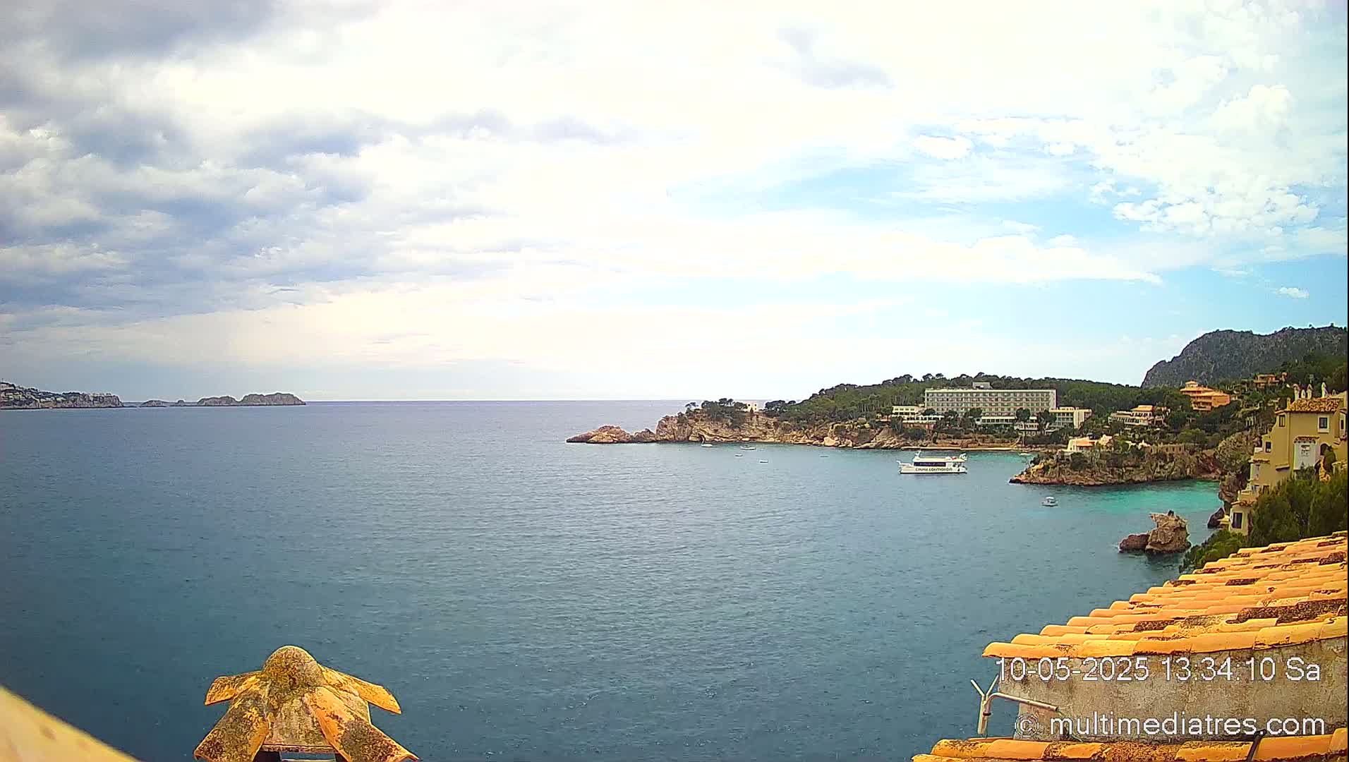 A partly cloudy day reveals a coastal scene with a calm blue sea, rocky shores, buildings, and a boat.