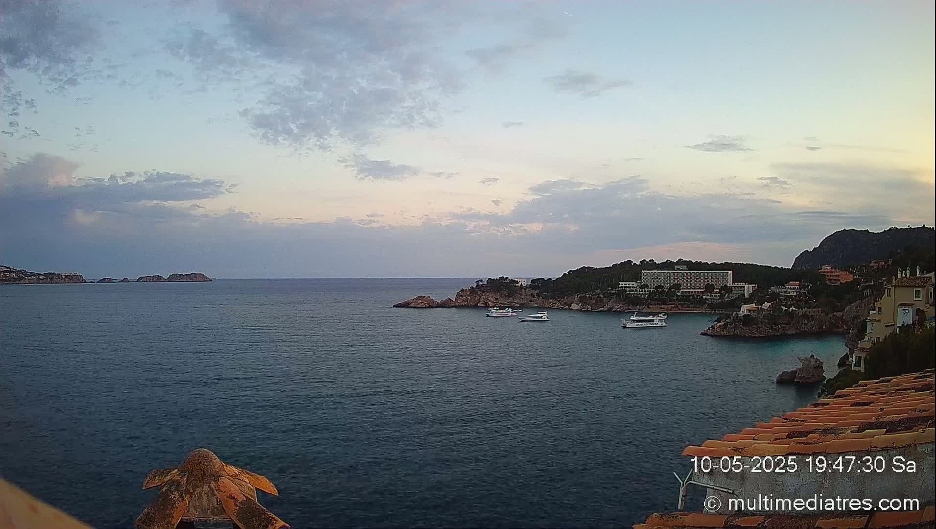 A calm sea with several boats near a coastline featuring buildings and rocky outcrops under a partly cloudy sky.