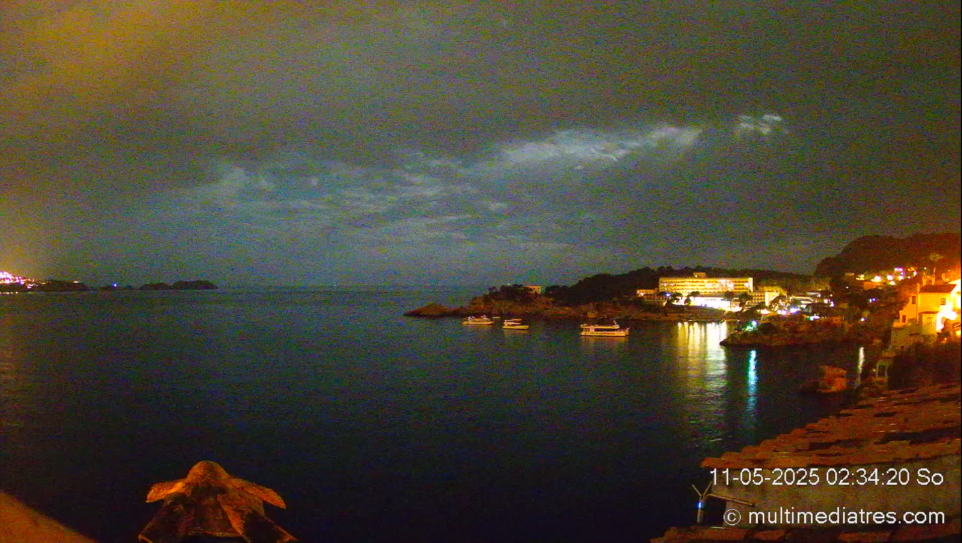 A nighttime coastal scene shows a dark body of water with several boats at a shore featuring buildings with illuminated windows under a cloudy sky.
