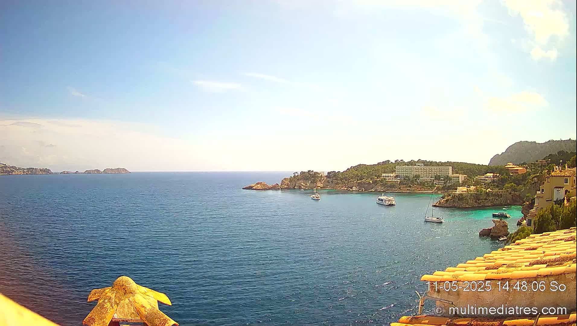 A sunny, coastal scene shows a calm blue sea with several boats, rocky shorelines with buildings, and distant islands under a clear sky.