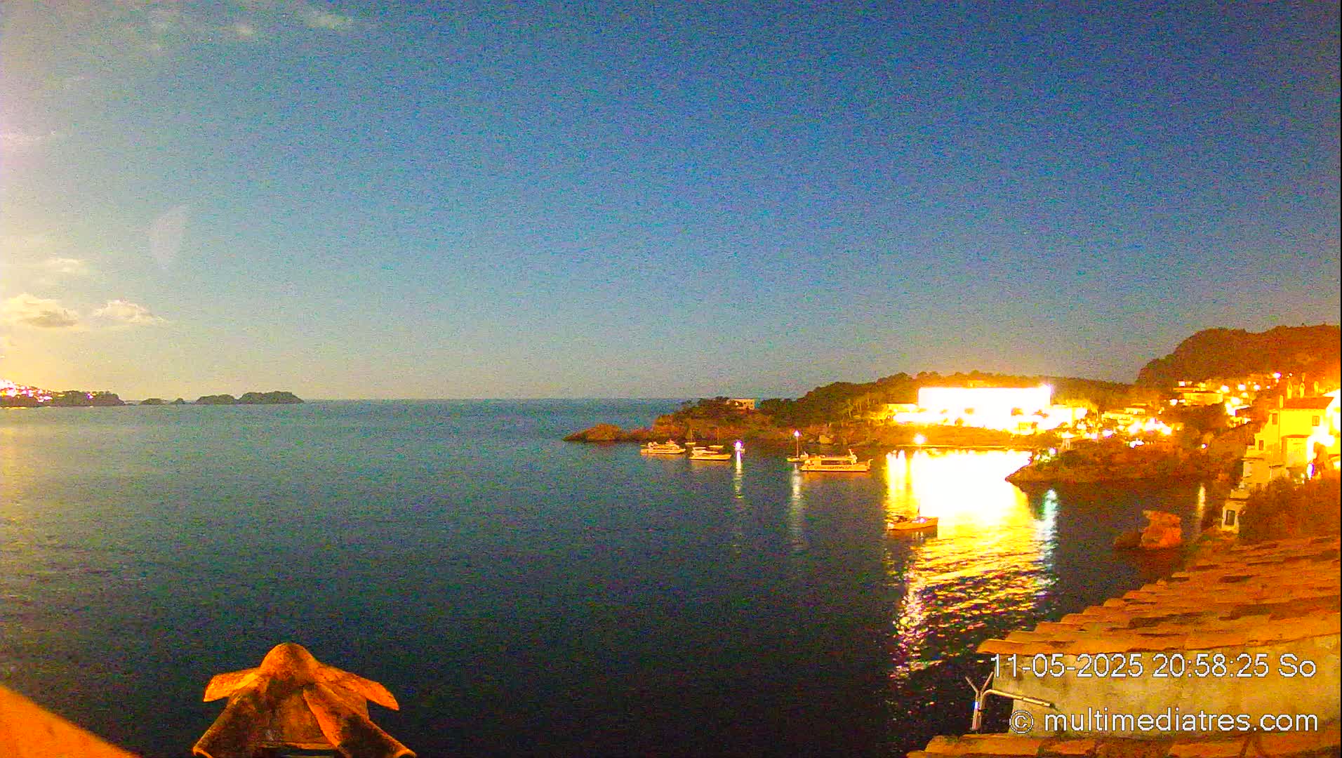 A nighttime view of a calm bay with boats, illuminated buildings on a hillside, and a clear, dark blue sky.