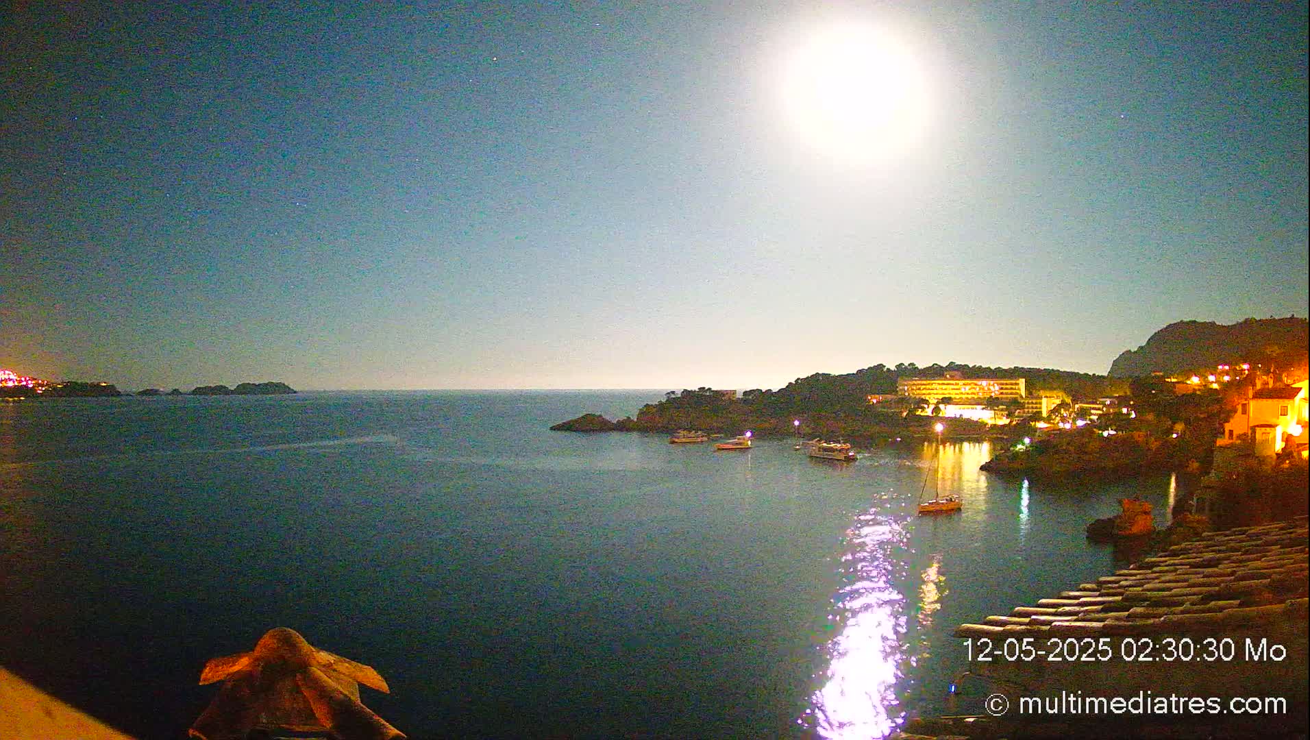 A bright moon illuminates a calm body of water at night, with a few boats in a harbor near a town nestled against a hillside.