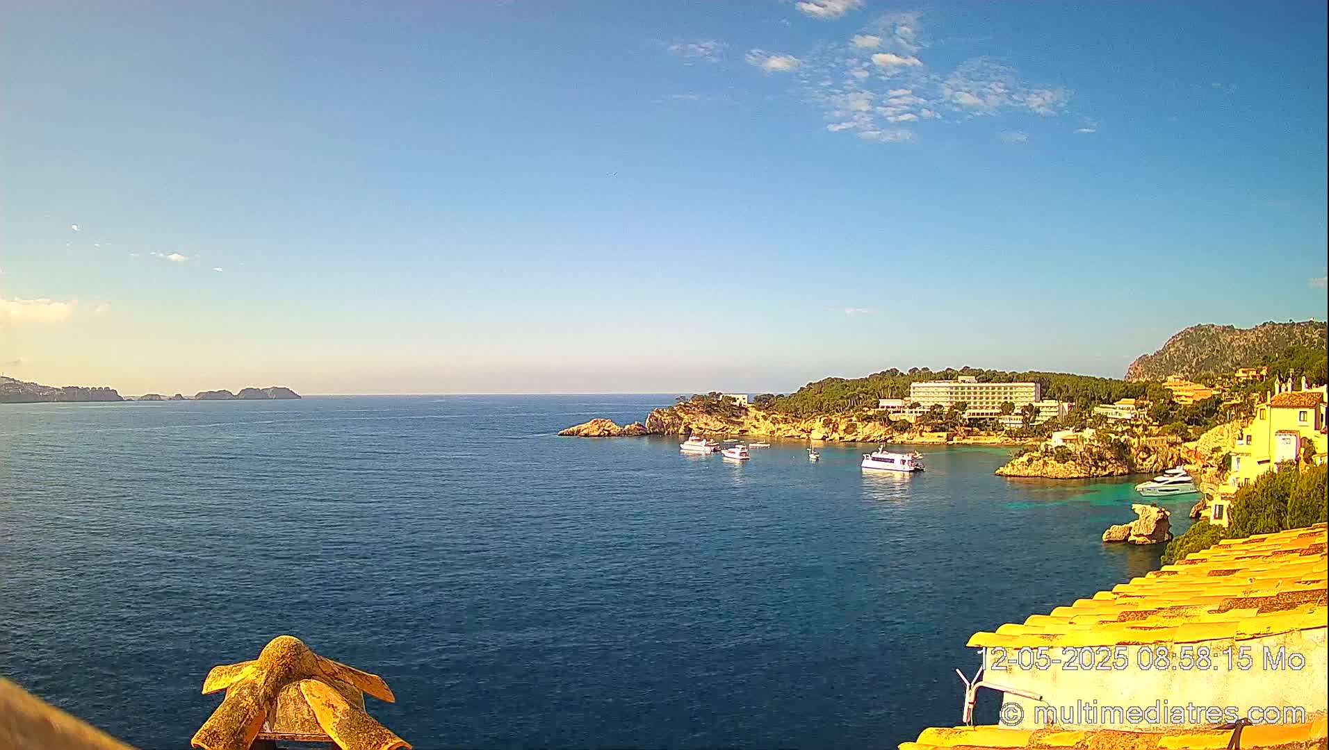 A calm, blue sea stretches out to a distant coastline under a mostly clear, sunny sky, with several boats anchored near a rocky shore lined with buildings and vegetation.