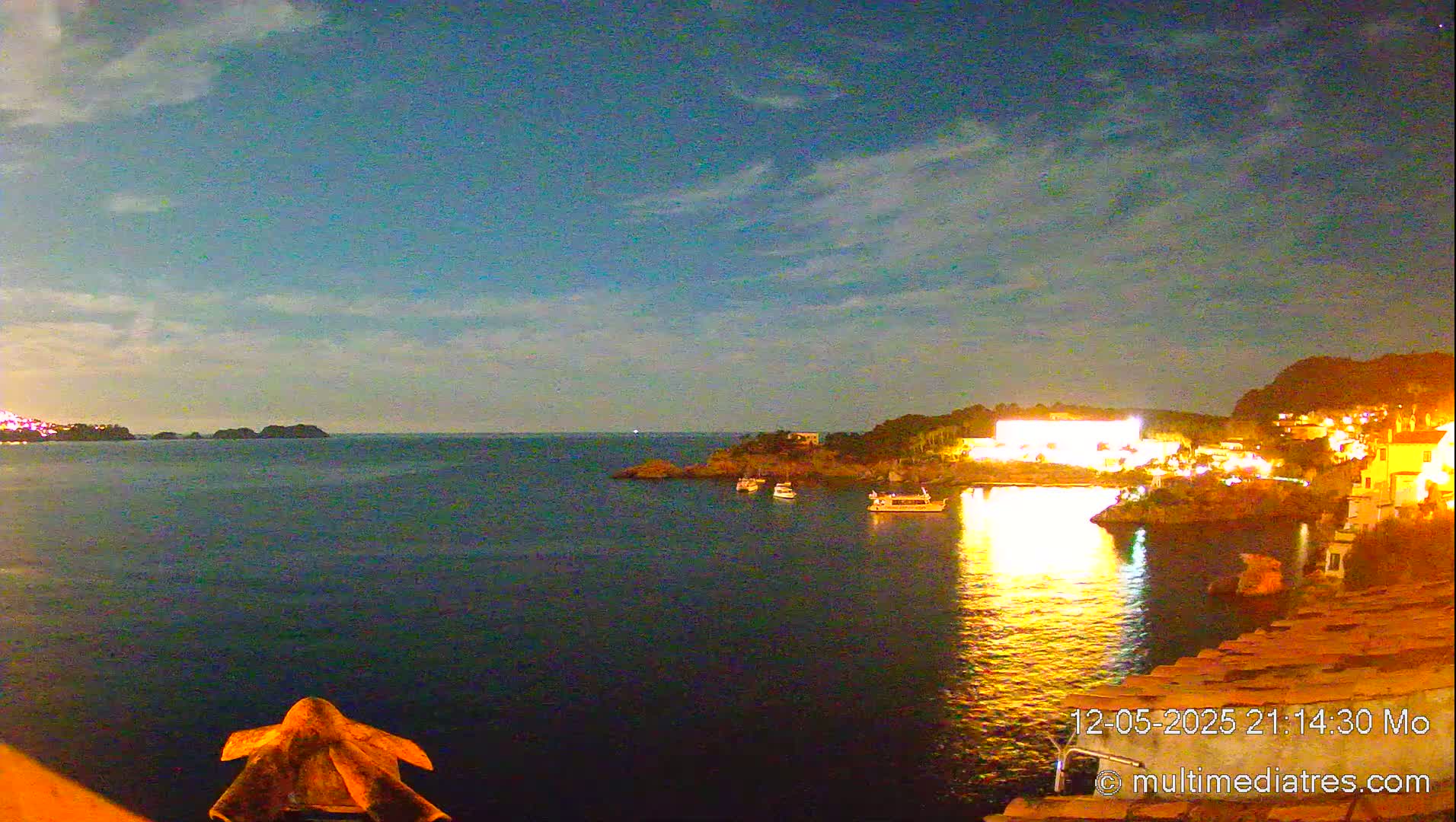 A nighttime coastal scene shows a calm body of water reflecting the lights of a village on a hillside under a partly cloudy sky.