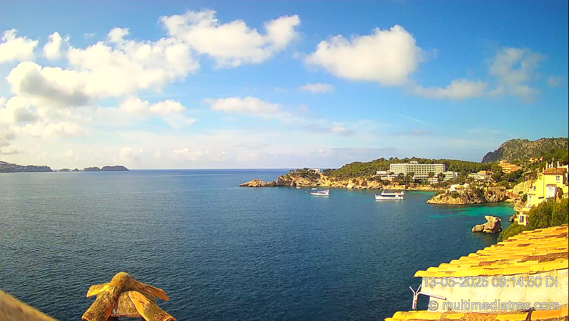 A partly cloudy blue sky overlooks a calm sea with a coastline featuring buildings, vegetation, and a few boats.