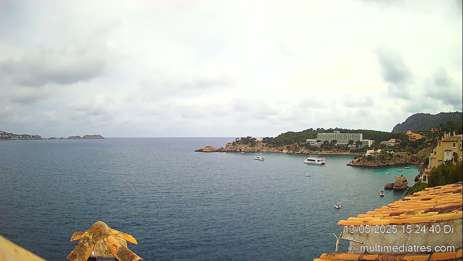 A partly cloudy day shows a tranquil coastal scene with a hotel and several boats in a calm bay, surrounded by rocky shores and distant islands.