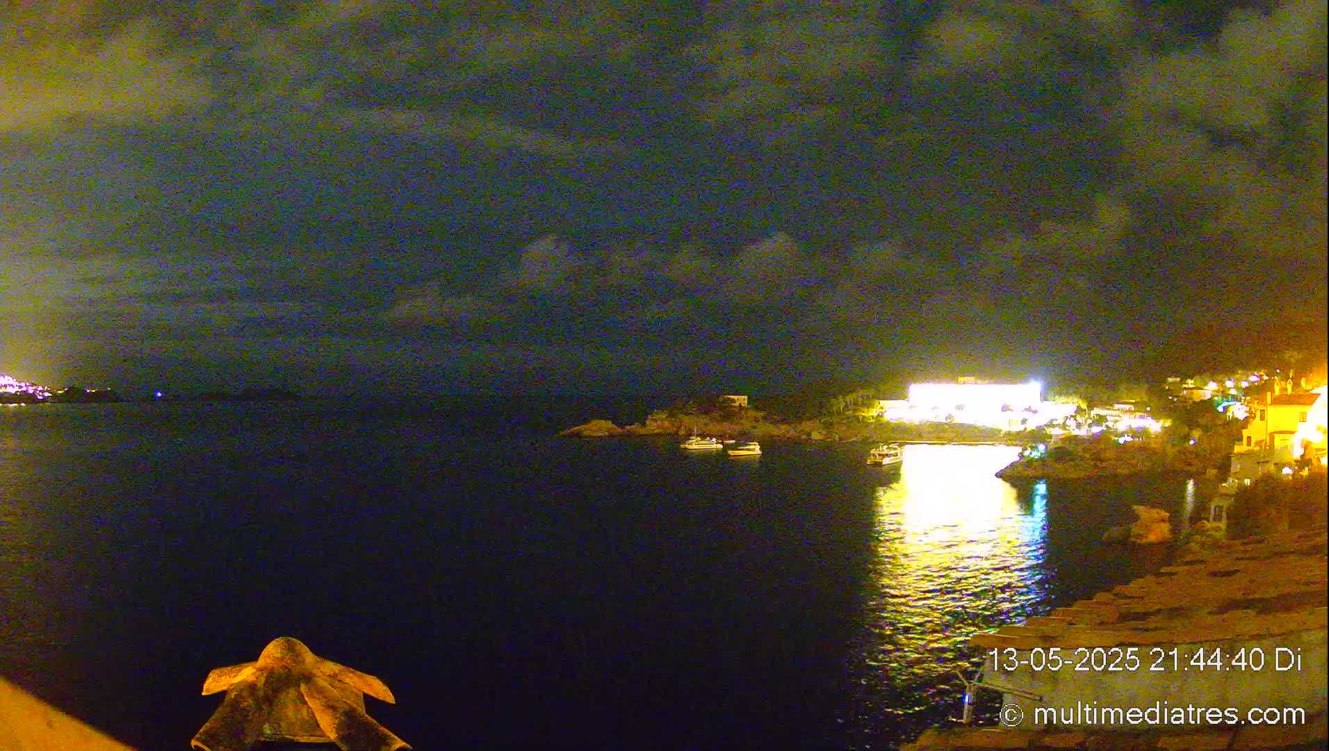 A nighttime view of a calm body of water with boats and a brightly lit shoreline under a cloudy sky.