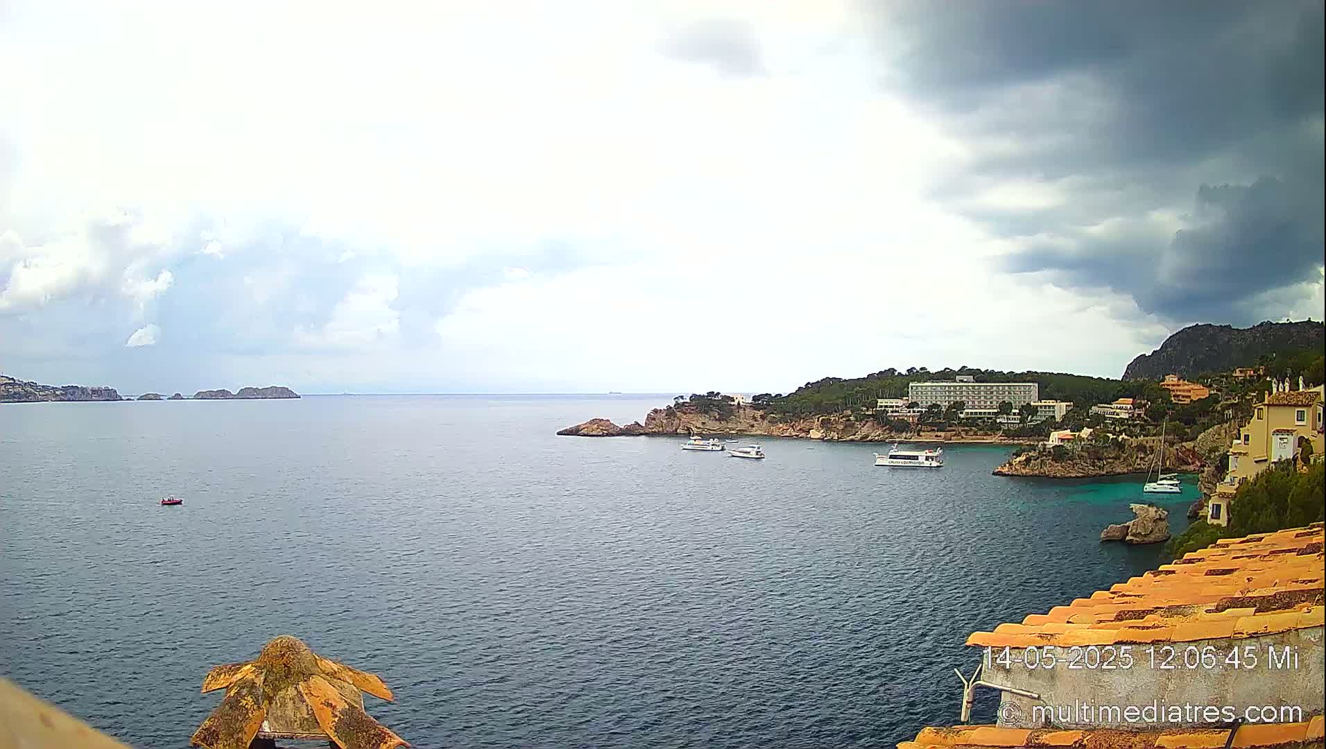 A partly cloudy, possibly stormy, ocean view shows several boats near a rocky coastline with buildings and vegetation.