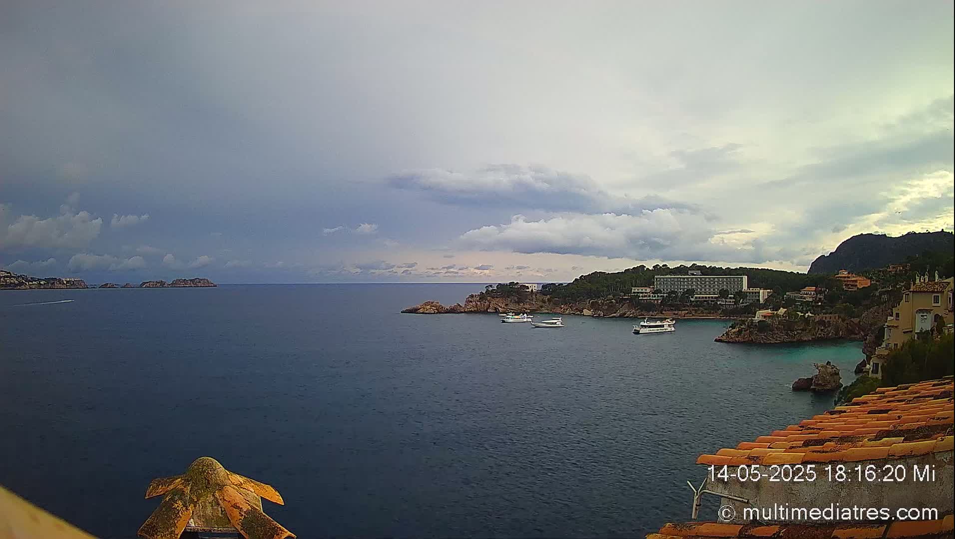 A partly cloudy sky overlooks a calm sea with several boats near a coastline featuring buildings and green hills.