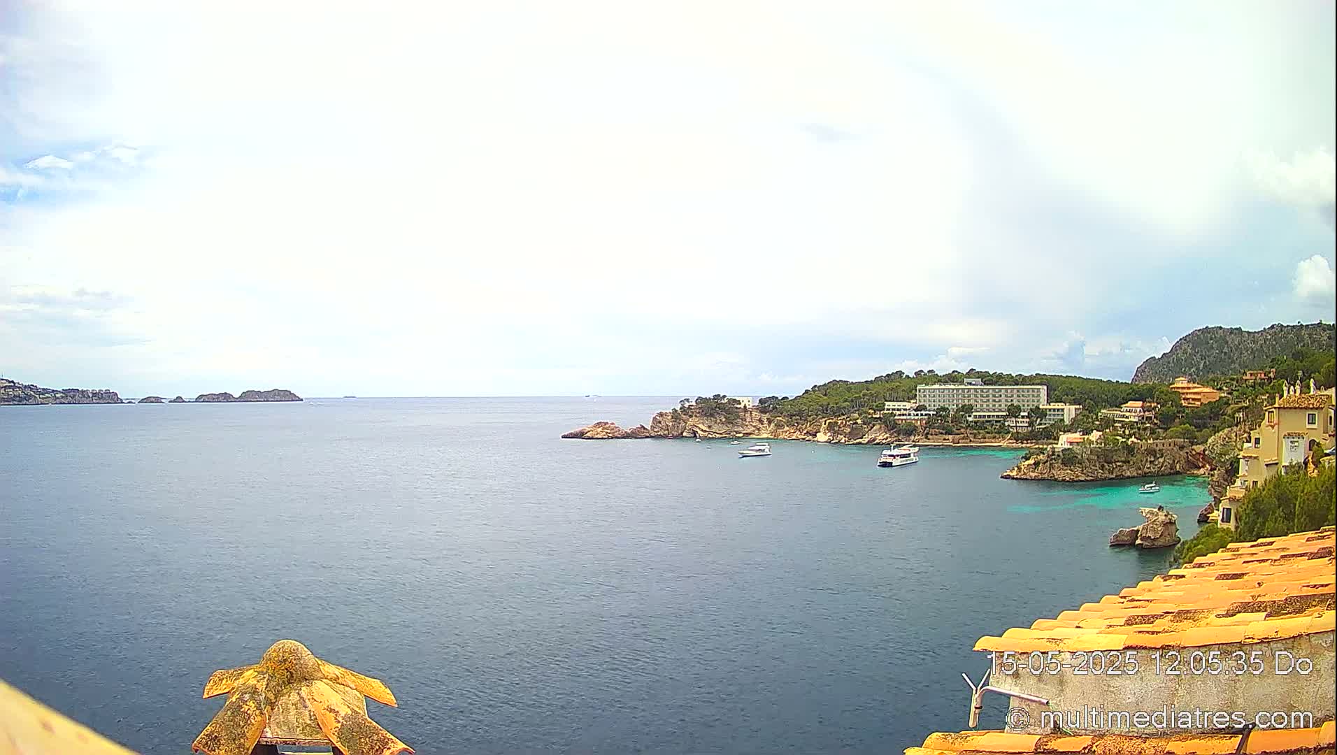 A tranquil coastal scene under a partly cloudy sky shows a calm sea with small boats, a rocky shoreline with buildings and vegetation, and distant islands.