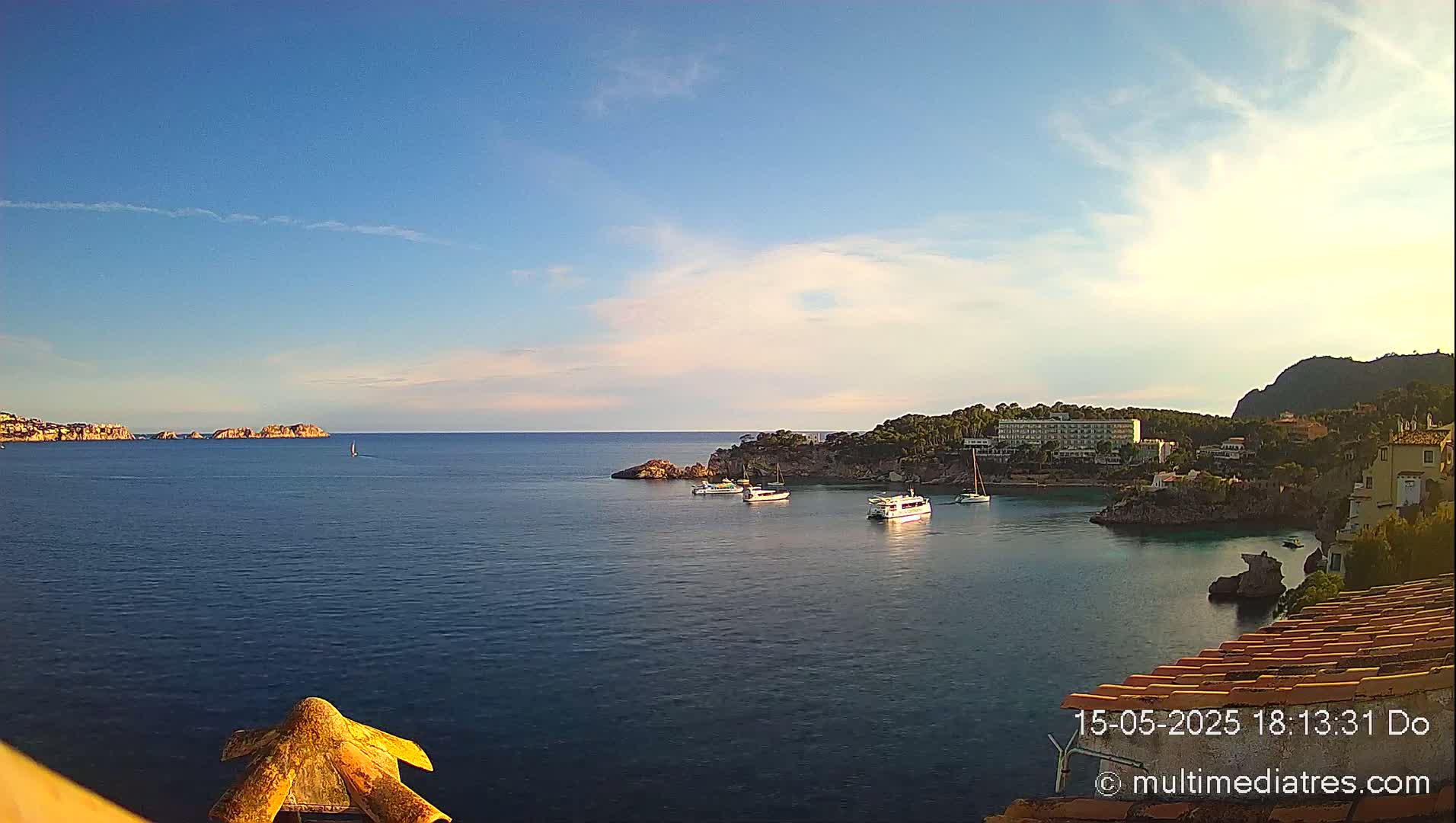 A tranquil coastal scene under a mostly clear, bright sky shows a calm bay with several boats, a hotel, and rocky headlands.