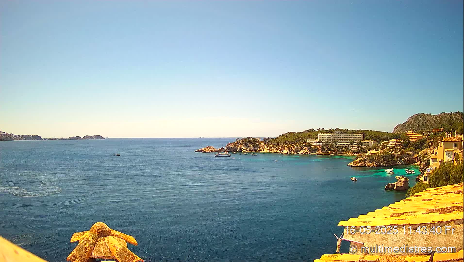 A sunny, clear day reveals a coastal scene of a calm blue sea with a few boats, rocky shores, and buildings nestled among green vegetation.