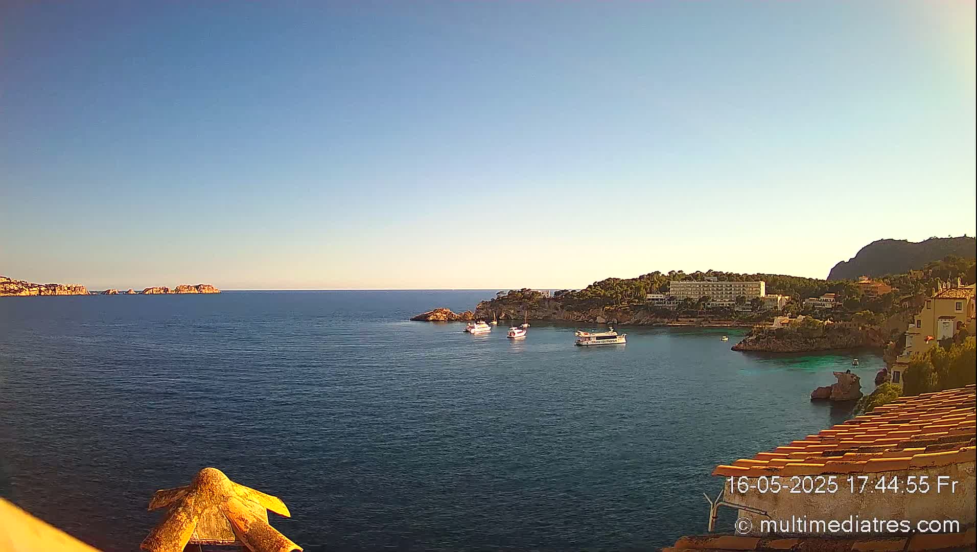 A clear, sunny day overlooks a calm bay with several boats, a coastline with buildings, and distant islands.