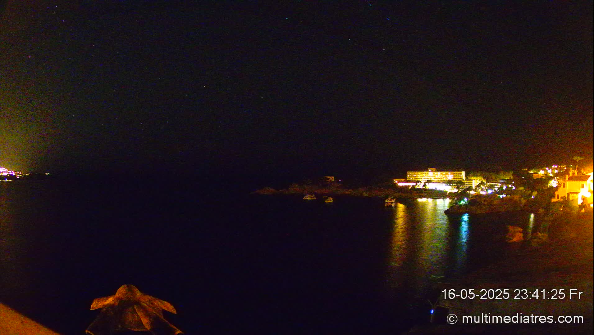 A nighttime view of a calm body of water reflecting the lights of a coastal town under a starry sky.