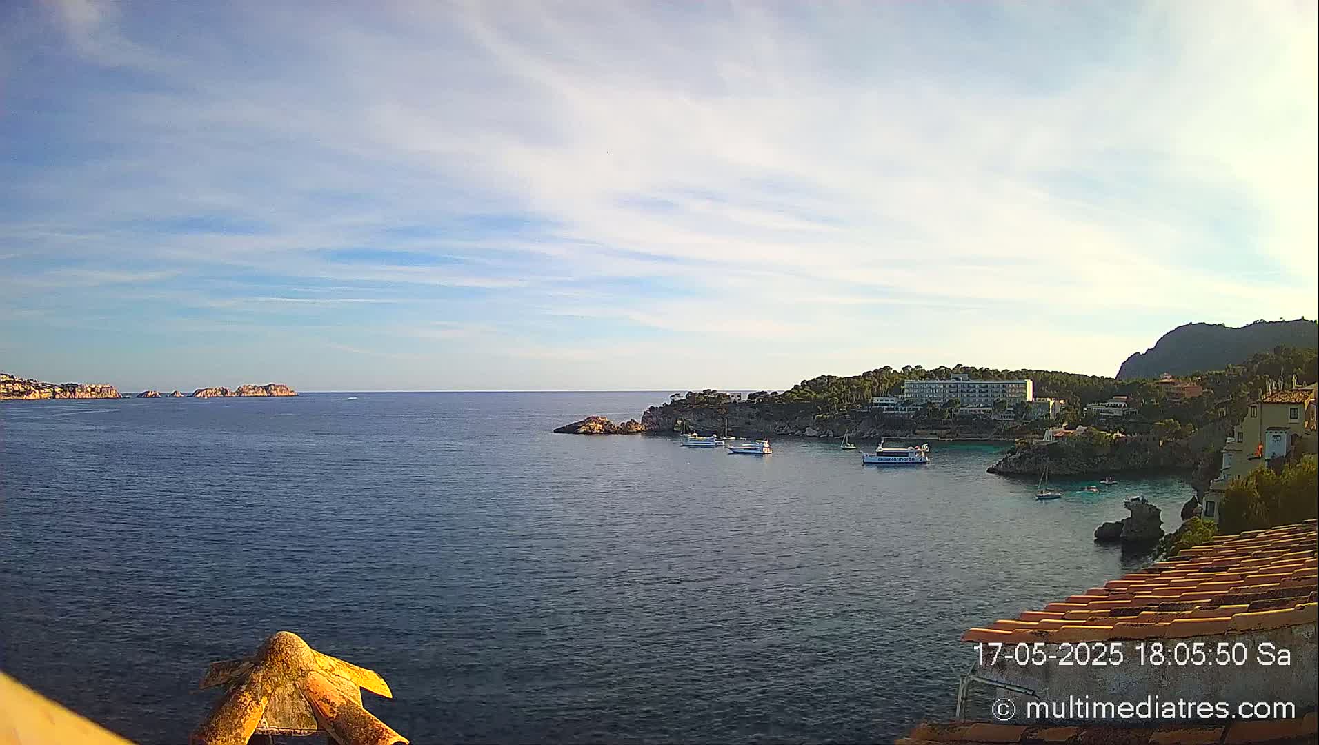 A calm, partly cloudy day reveals a picturesque coastal scene with several boats in a sheltered bay, nestled between rocky outcrops and buildings on a hillside.