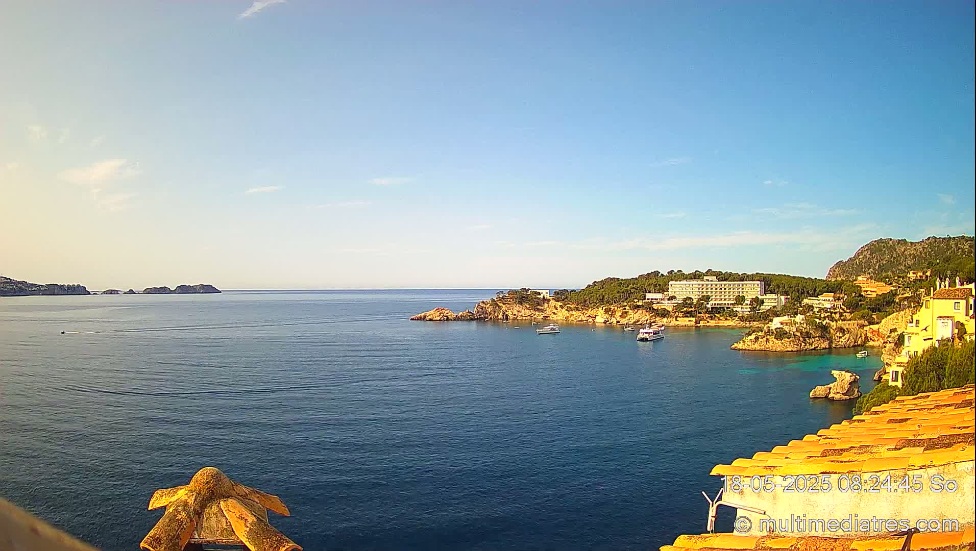 A calm, clear day reveals a coastal scene with a hotel and buildings nestled on a rocky hillside overlooking a bay with several boats.