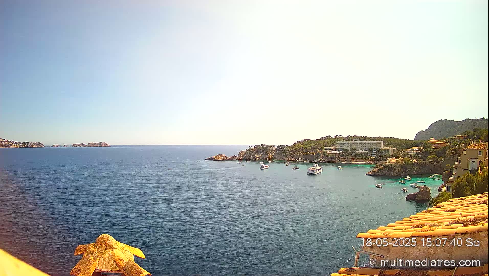 A tranquil, sunny coastal scene shows a calm blue sea with several boats near a rocky shoreline and buildings, under a clear sky.