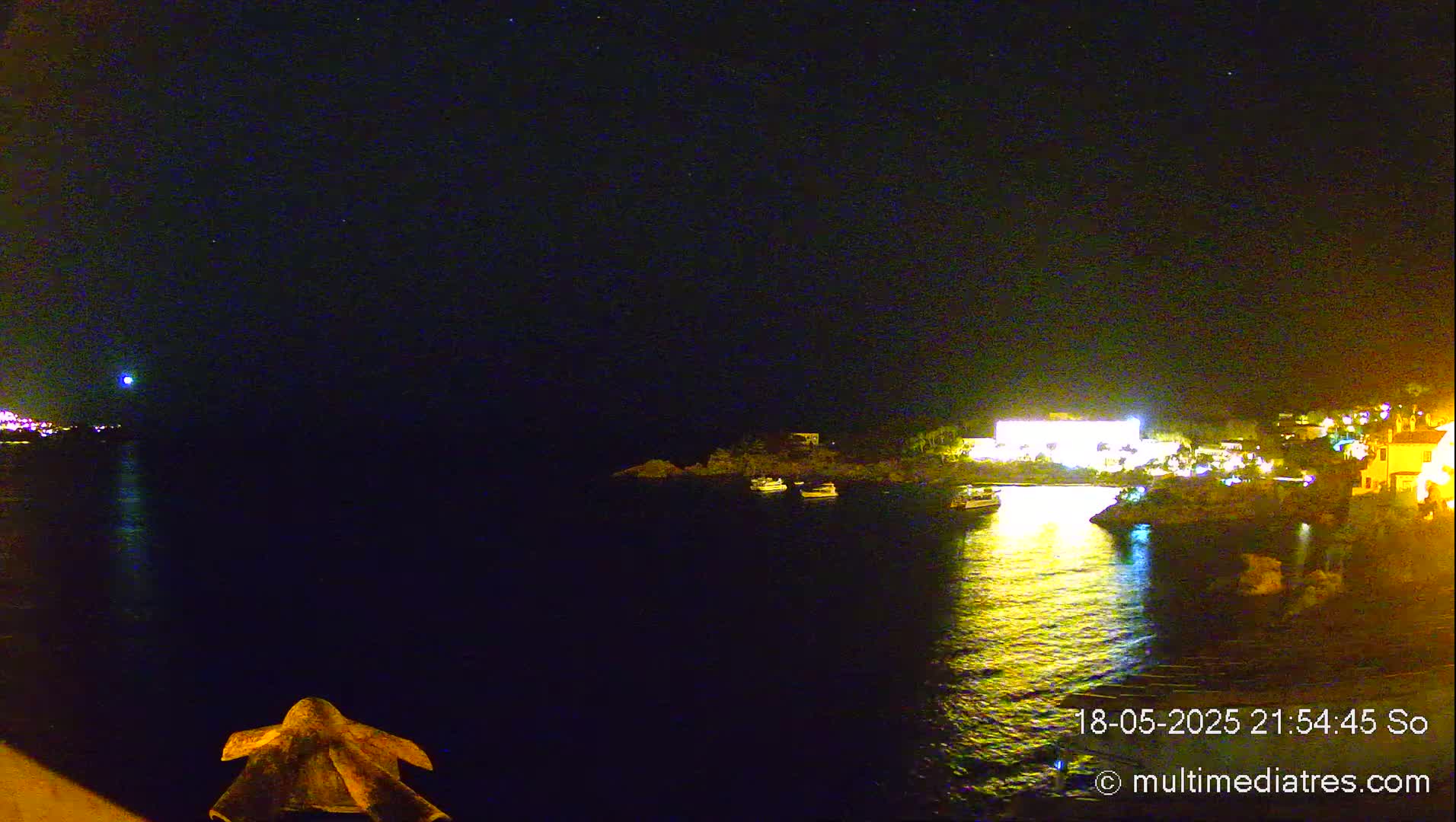 A nighttime view of a calm body of water with illuminated buildings and boats on the shore under a clear, starry sky.