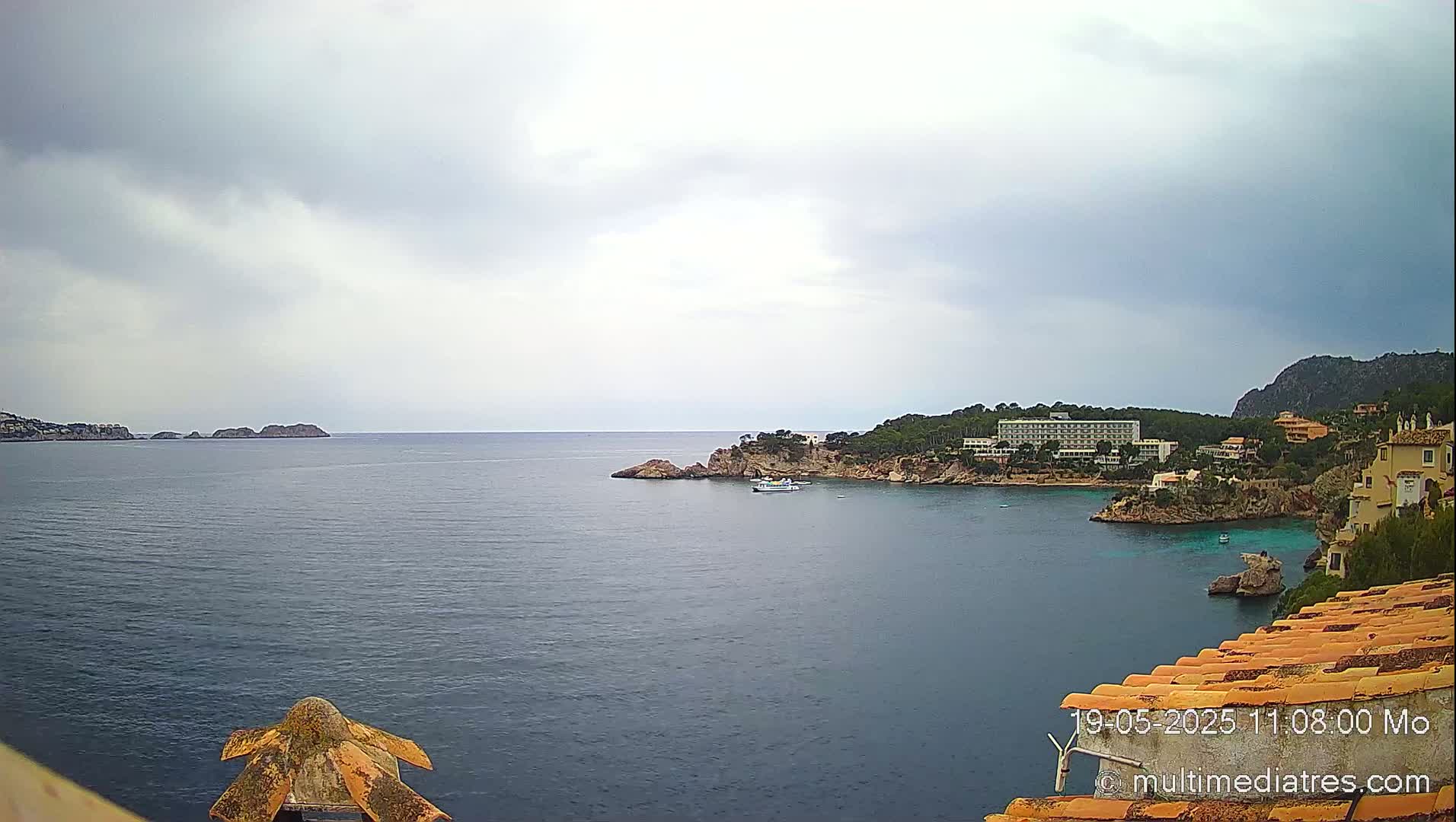 A coastal scene under an overcast sky shows a calm sea with rocky shores, buildings, and a small boat.