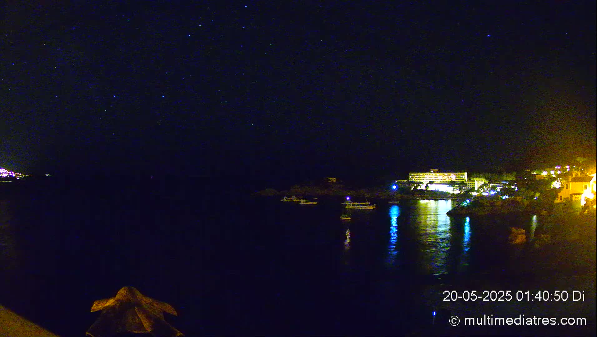 A nighttime view of a calm body of water with several boats, a brightly lit building on the opposite shore, and a starry sky above.