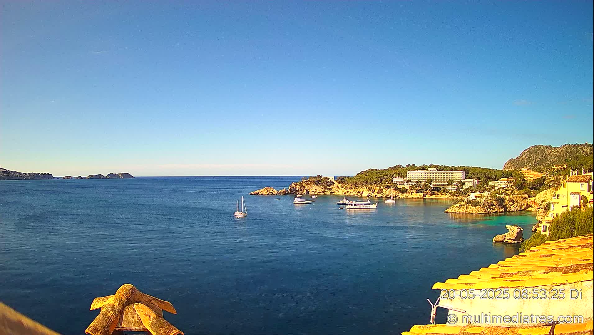 A calm, blue ocean bay with several boats, a hotel, and buildings on a rocky shore under a clear, bright blue sky.