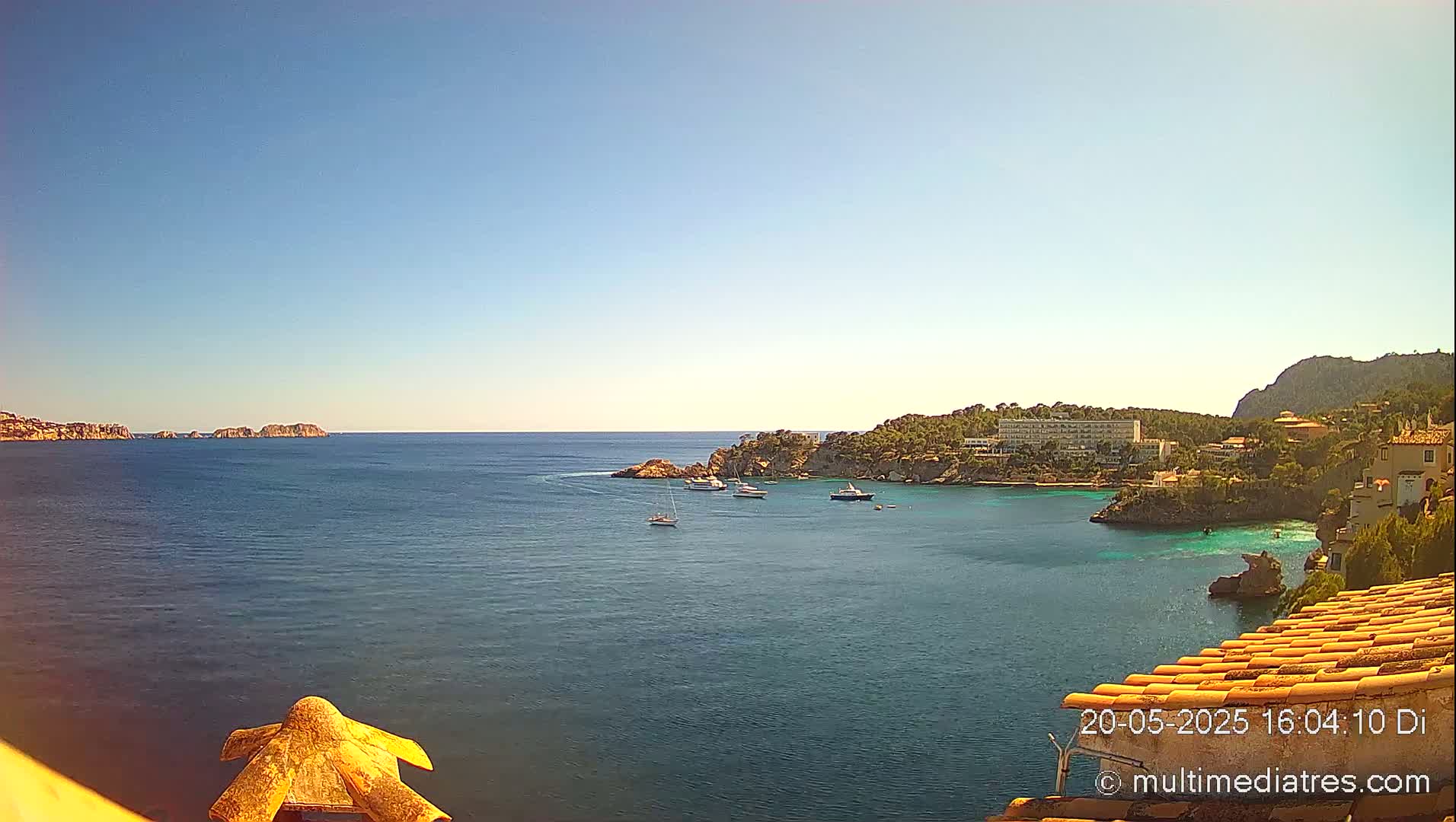 A tranquil ocean bay on a sunny day, with several boats anchored near a rocky shoreline featuring buildings and vegetation.