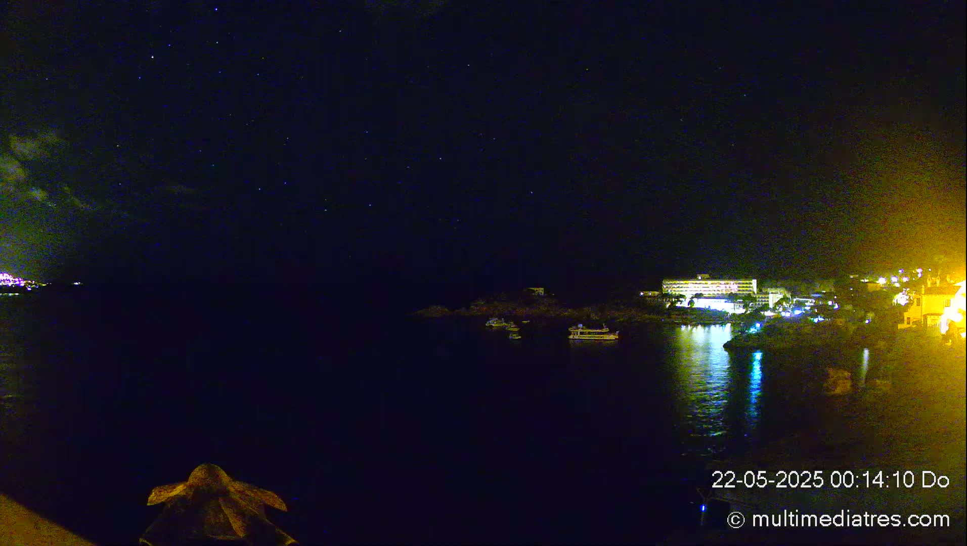 A nighttime view of a calm body of water with several boats and a lit-up building on the opposite shore under a starry sky.