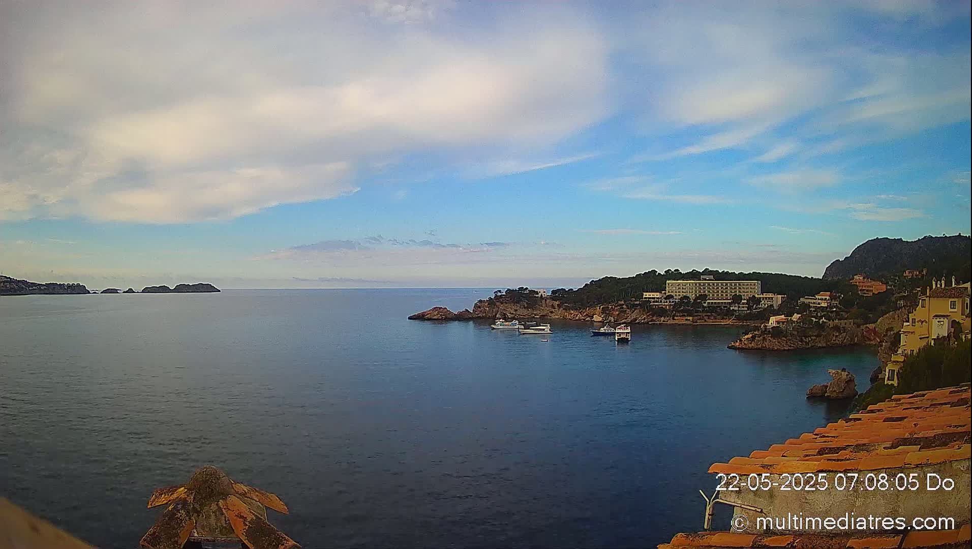 A partly cloudy blue sky overlooks a calm bay with several boats, a hotel, and buildings nestled along rocky coastlines.