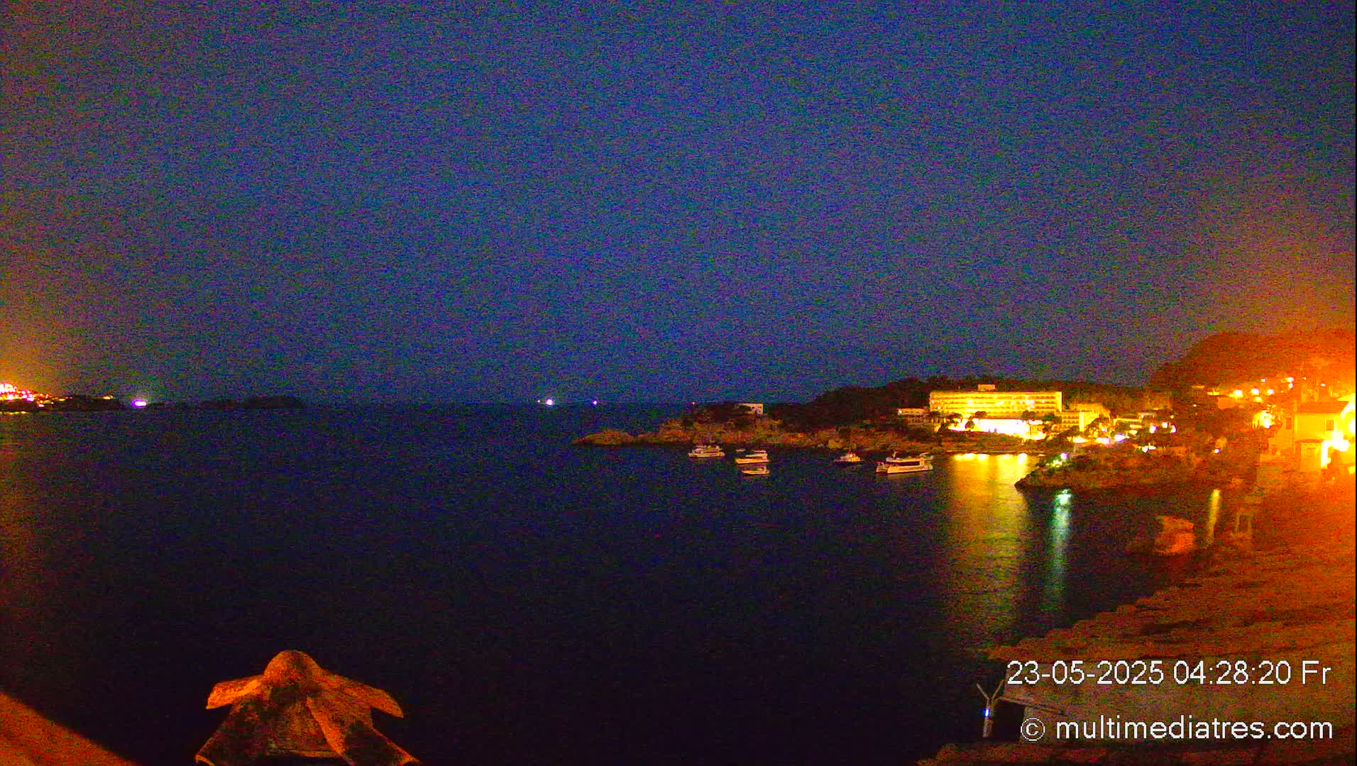 A nighttime coastal scene shows a dark body of water with several boats and a brightly lit hotel and town on the far shore under a clear, dark blue sky.