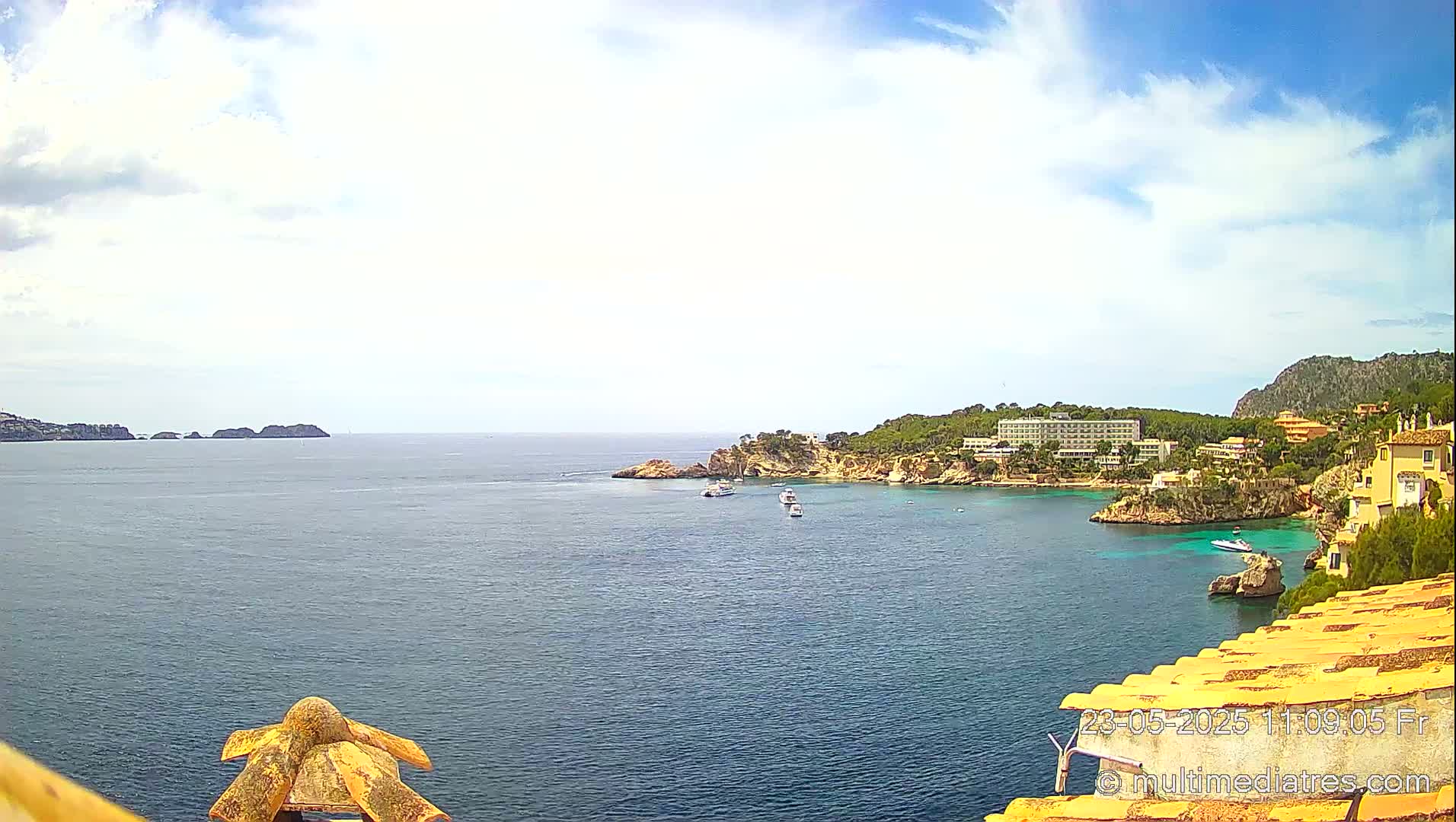 A partly cloudy day shows a coastal scene with a calm sea, several boats, and buildings on a rocky shoreline.