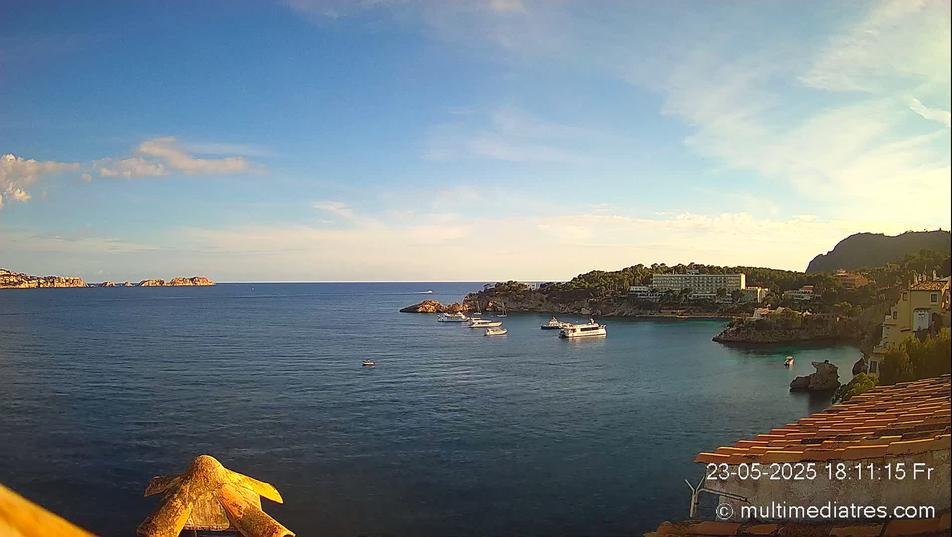 A calm, clear day reveals a coastal scene with several boats in a bay, surrounded by green hills and buildings, with a distant island visible on the horizon.