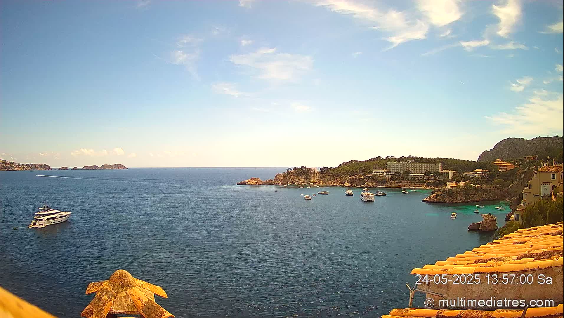 A yacht and several smaller boats are in a calm, blue ocean cove near a rocky shoreline with buildings and vegetation under a mostly sunny sky.