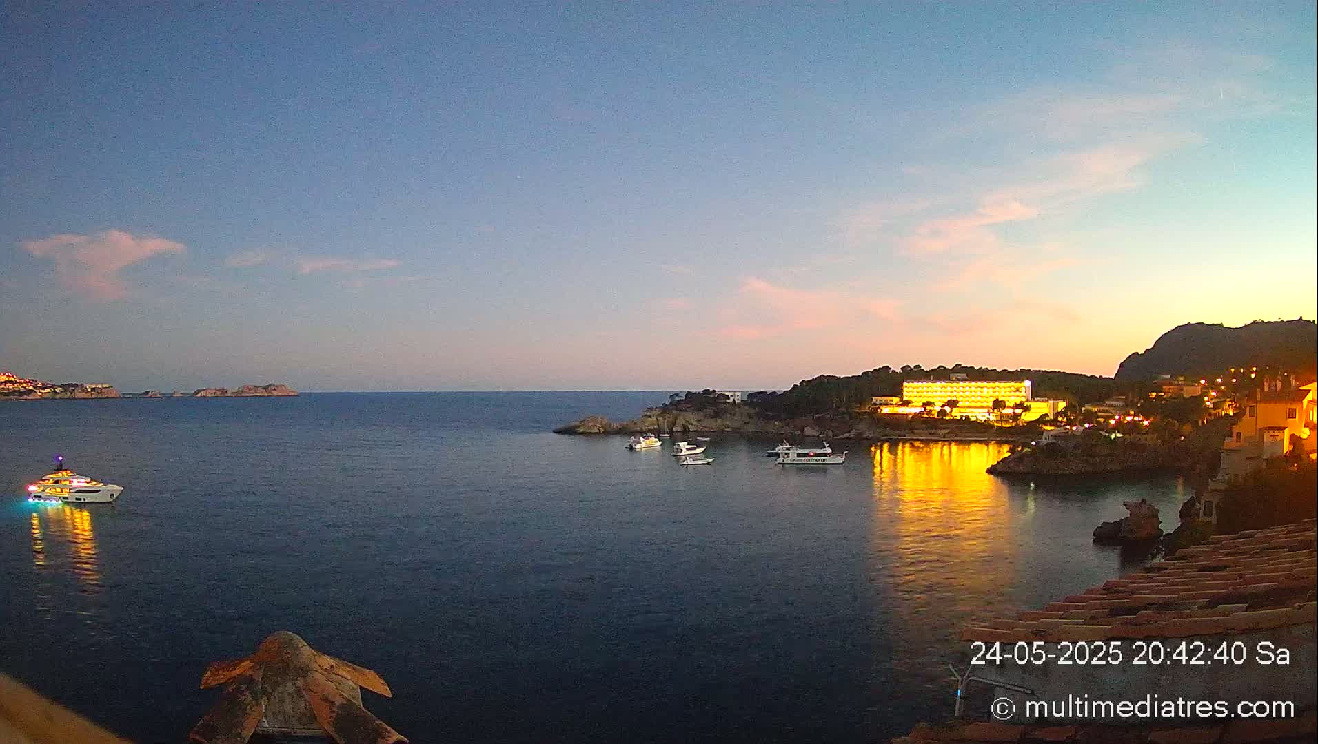 A calm twilight sea reflects the lights of a coastal hotel and town, with several boats anchored in the bay.