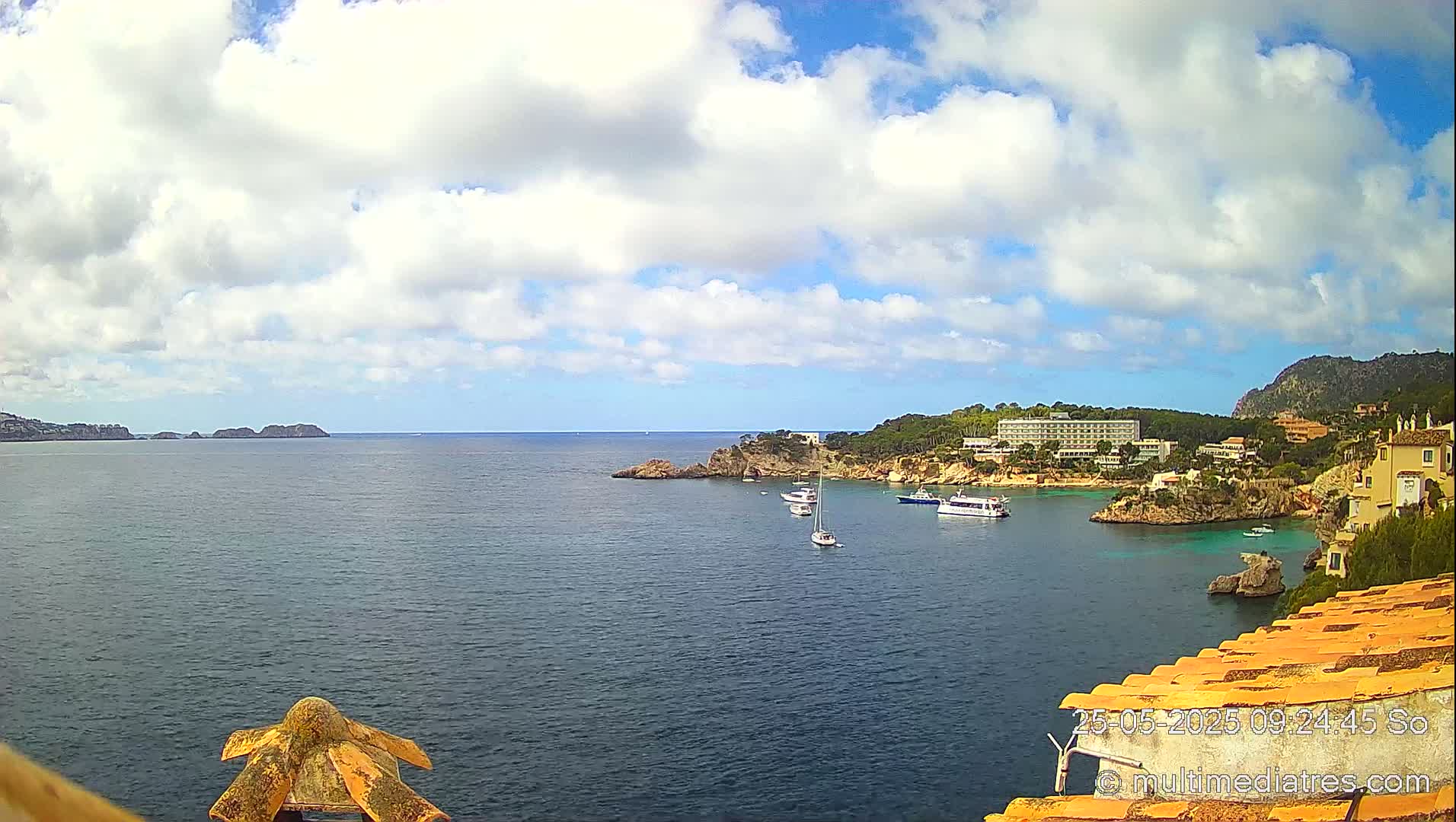 A partly cloudy day shows a calm bay with several boats anchored near a rocky shore and buildings, including a large hotel.