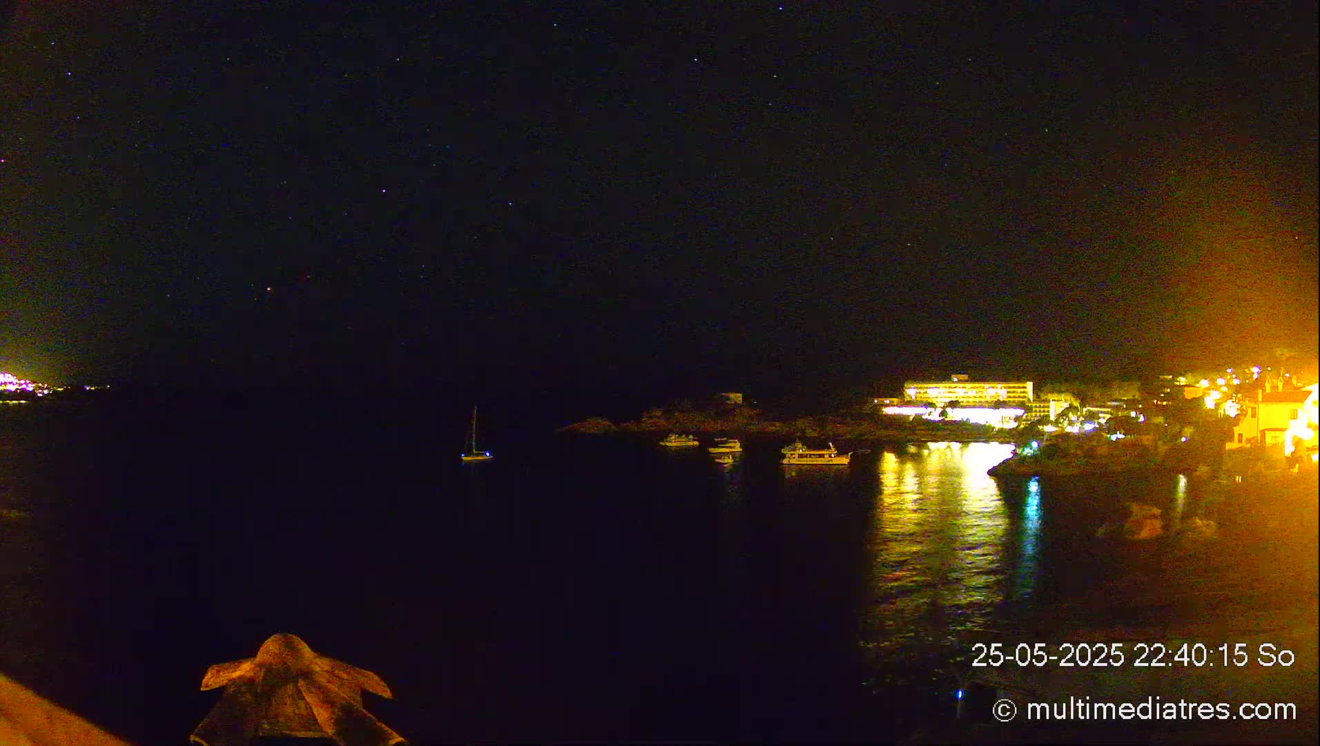 A nighttime view of a calm body of water reflecting the lights of a coastal town and a large building under a clear, starry sky.