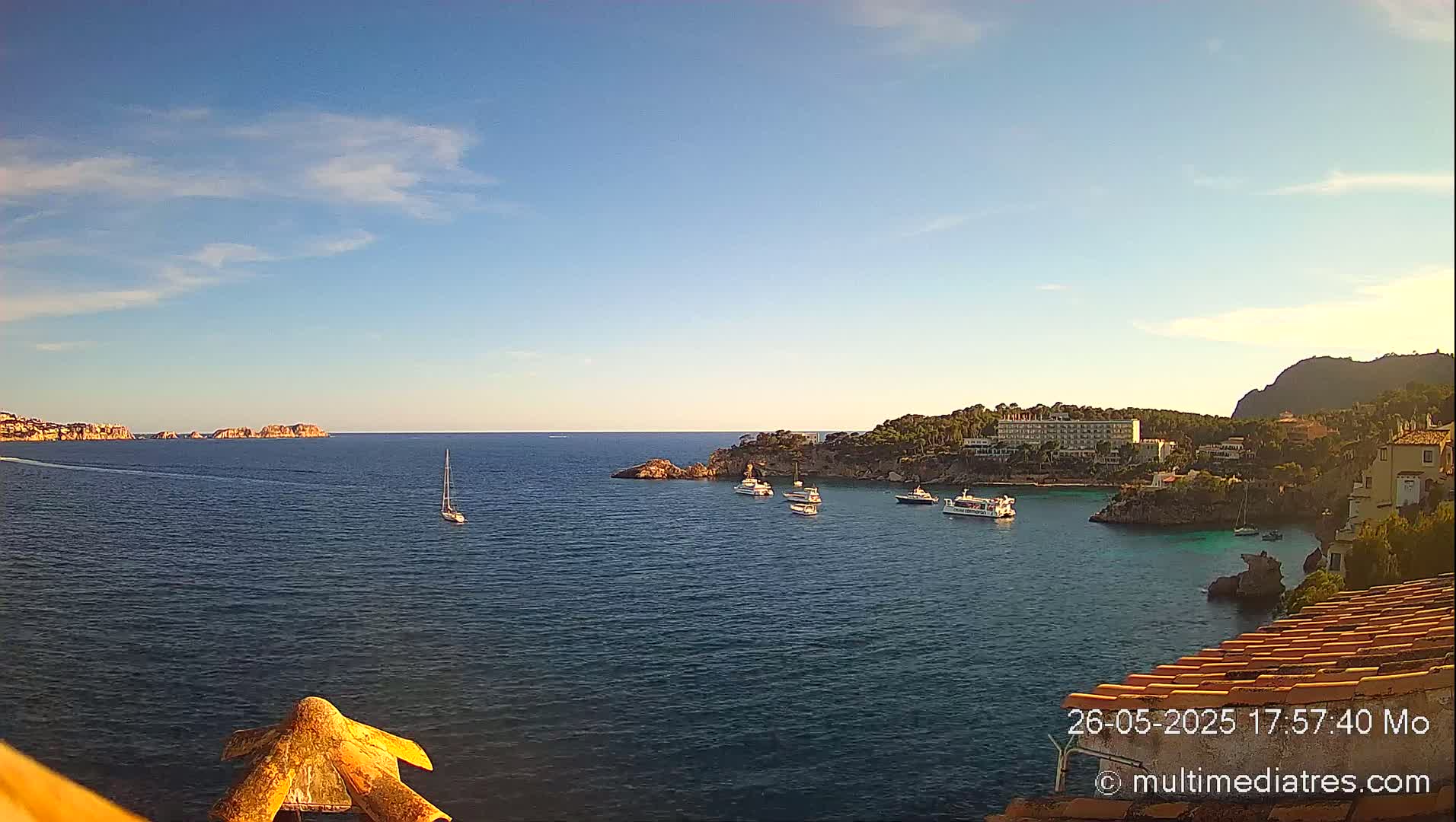 A tranquil coastal scene under a clear, sunny sky shows a calm sea with several boats anchored near a rocky shoreline and buildings.