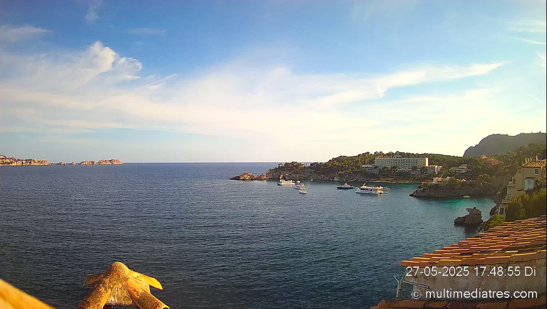 A tranquil ocean cove with several boats, a hotel, and buildings nestled along the rocky shoreline under a mostly clear, sunny sky.