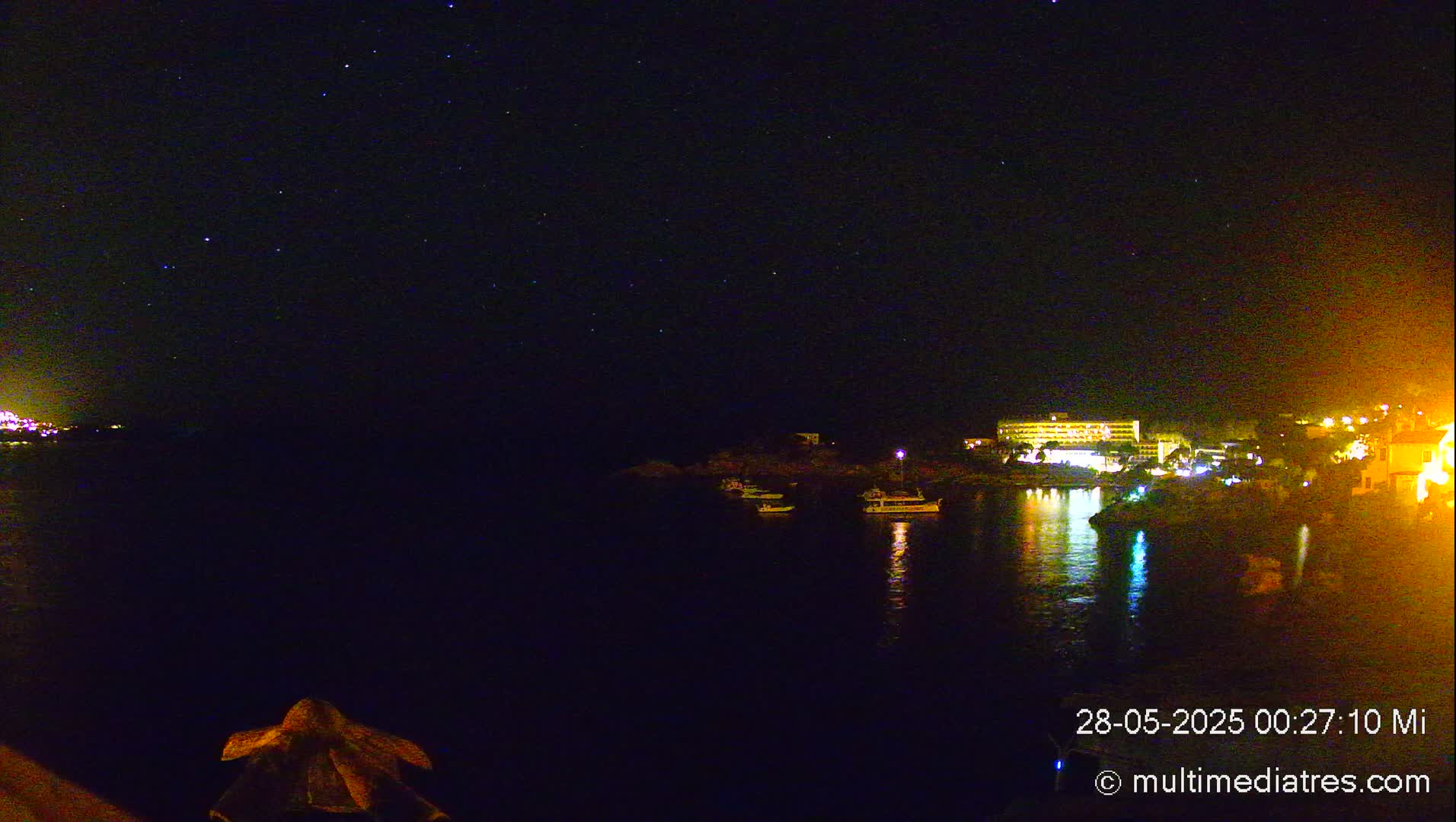 A nighttime view of a calm body of water with a few boats and a brightly lit building on the shore under a starry sky.