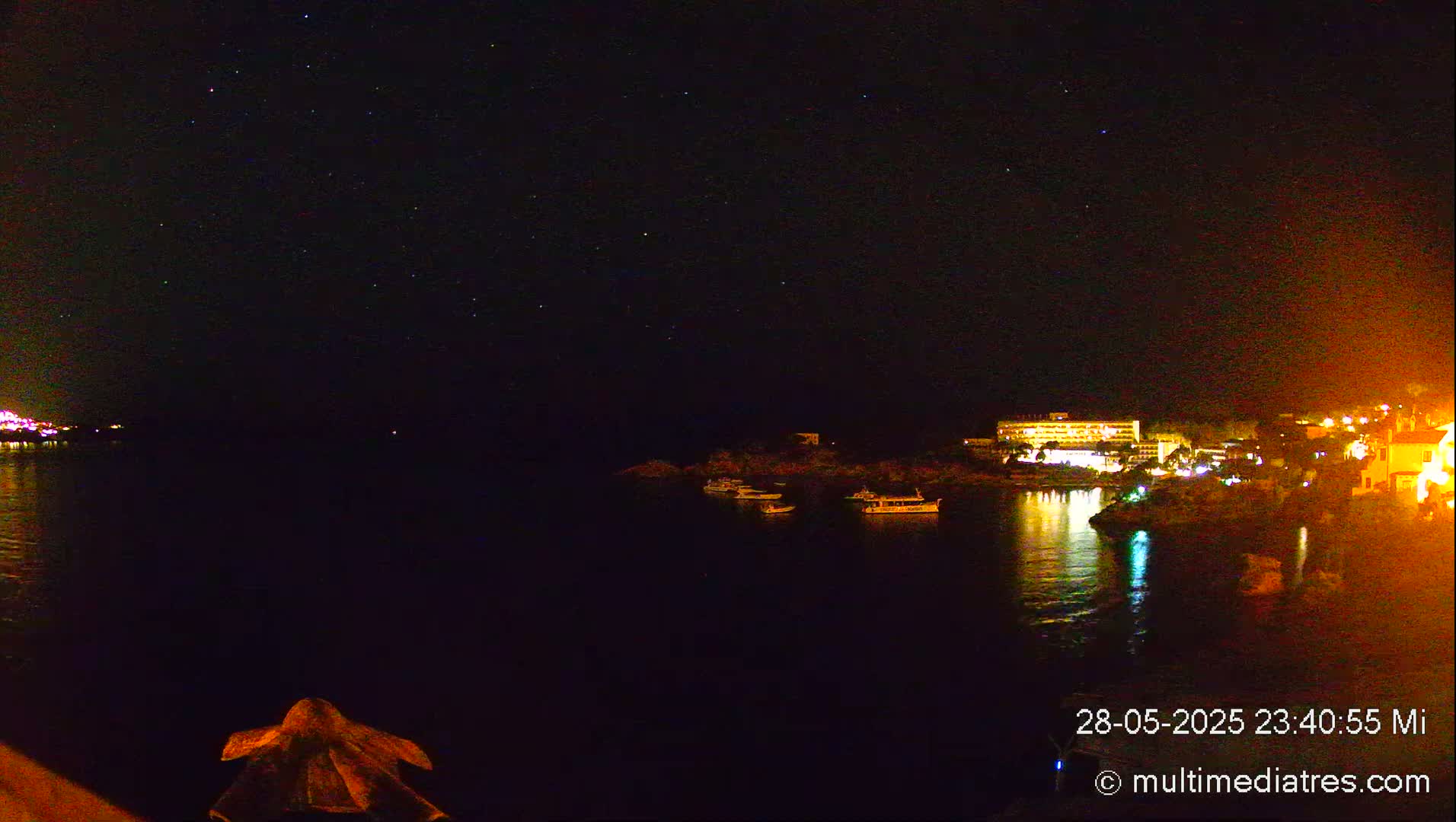 A nighttime view of a calm body of water with a few boats, illuminated buildings on the shore, and a starry sky above.
