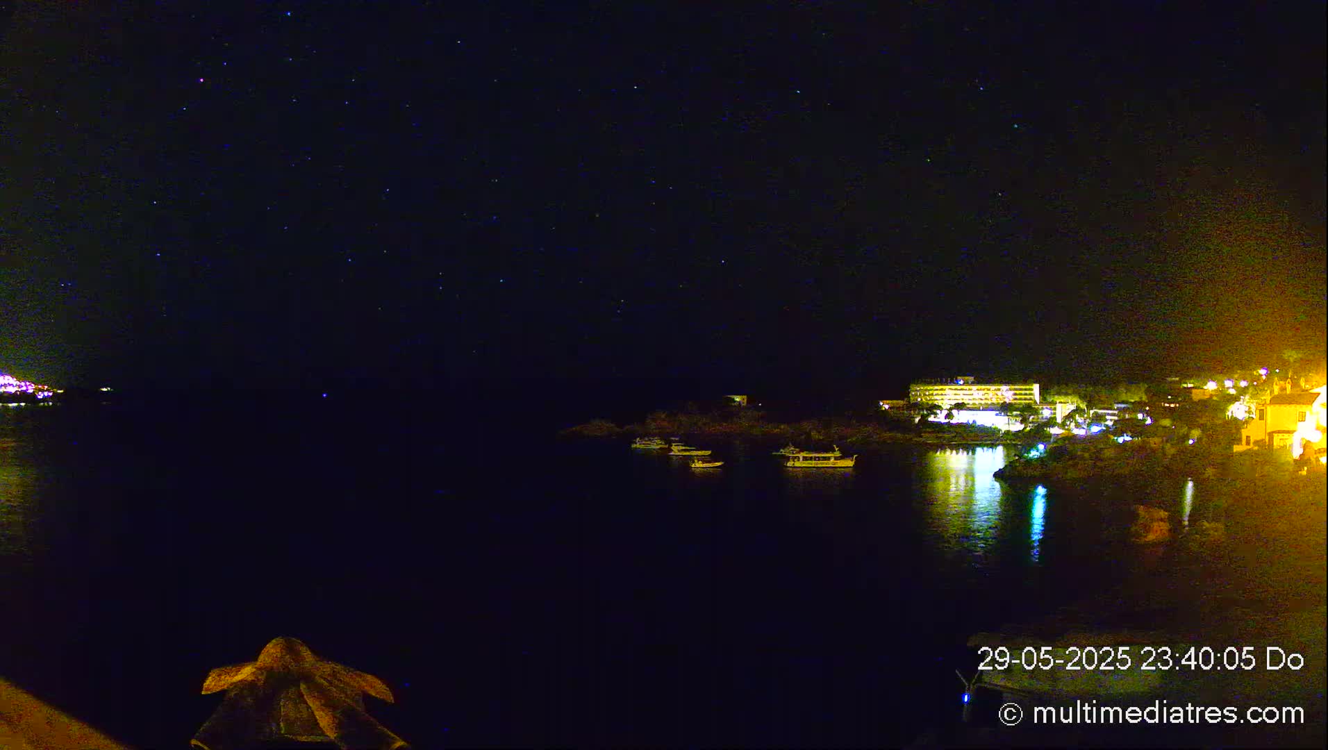 A nighttime view of a calm body of water with several boats, a lit-up shoreline featuring buildings, and a starry sky above.