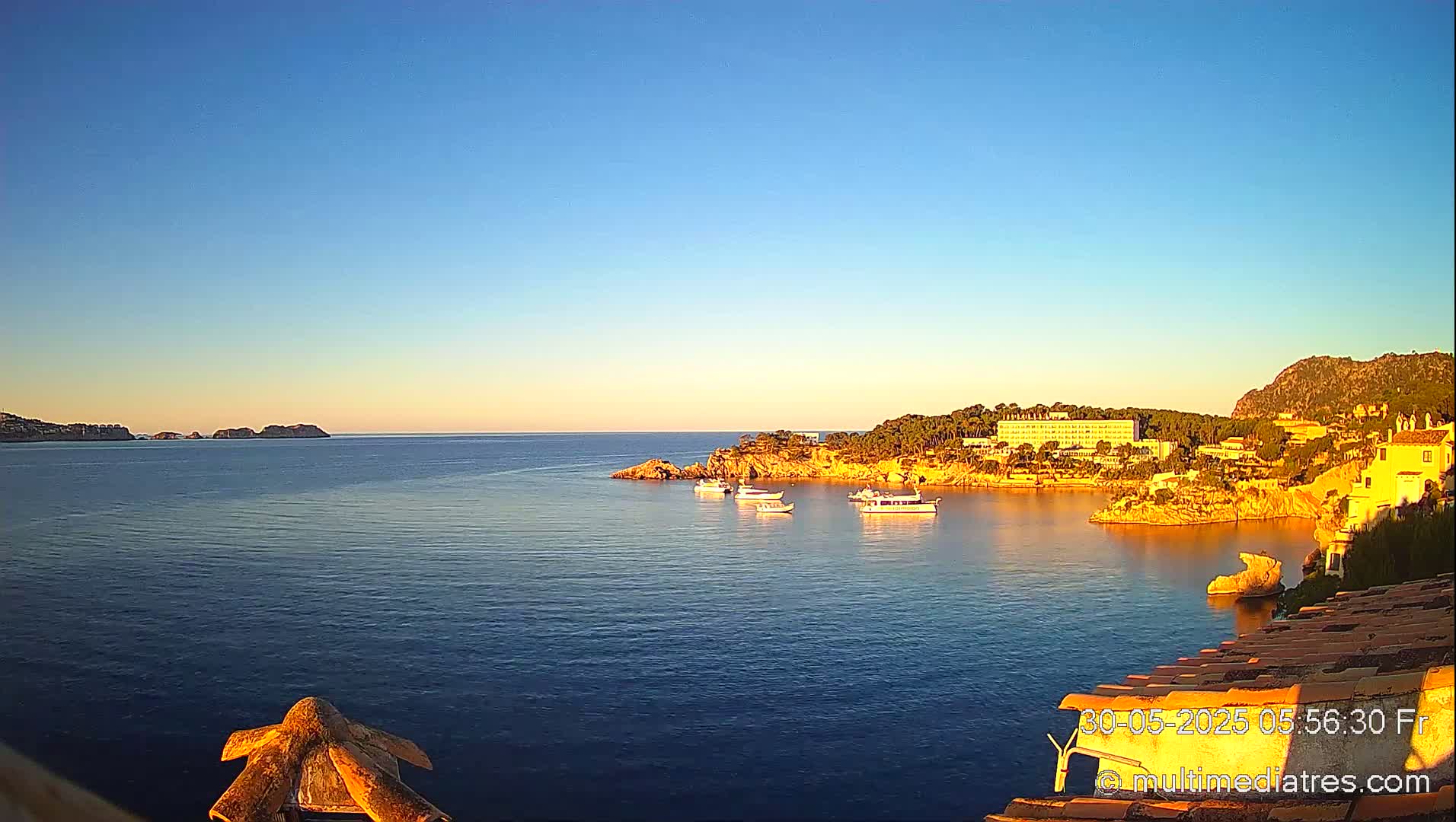 A calm, clear blue sea under a bright, mostly clear sky shows several small boats moored near a rocky coastline with buildings and trees.