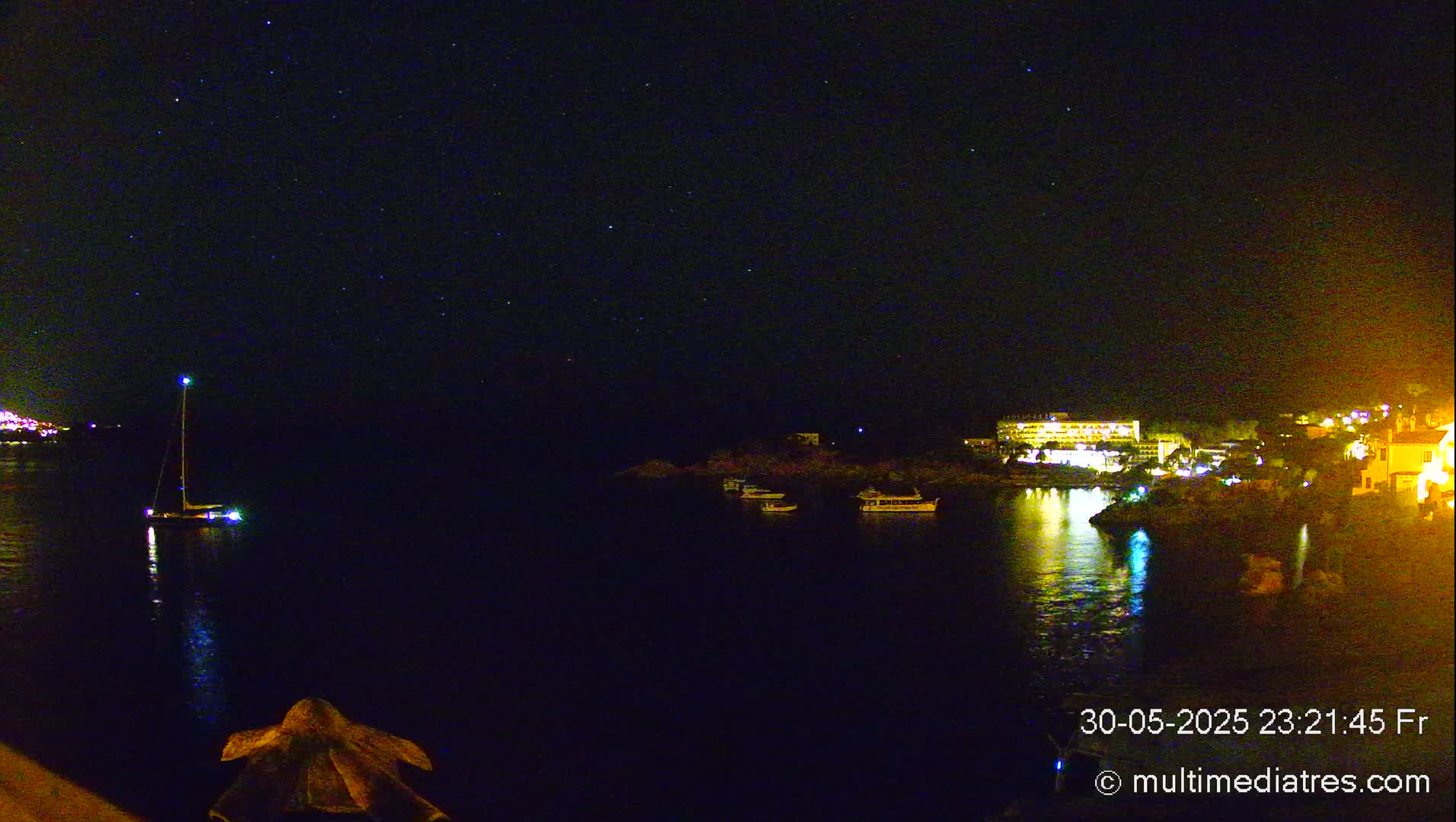 A nighttime view of a calm body of water with a sailboat and several other boats, a lit-up shoreline with buildings, and a starry sky above.